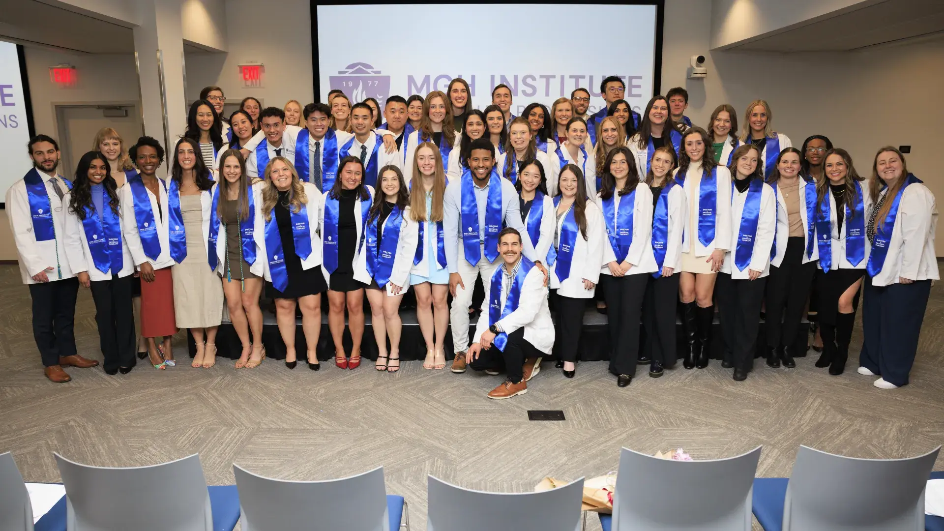 A large group poses for a photo wearing white coats and blue sashes around their necks