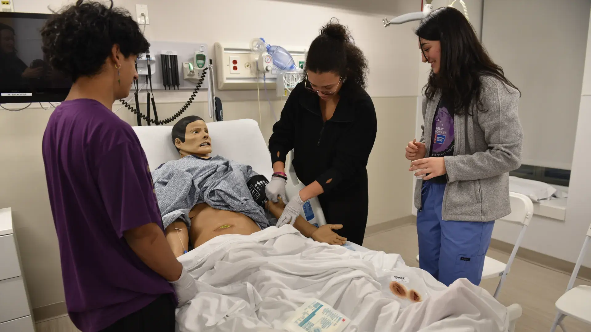 A woman works on a manikin in a hospital bed while another woman looks on next to her and a man watches from across the bed