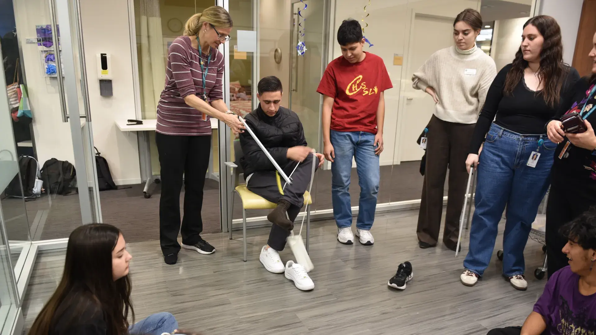 A person sits and finishes putting on a sock while a person stands next to him using a tool to grasp the sock