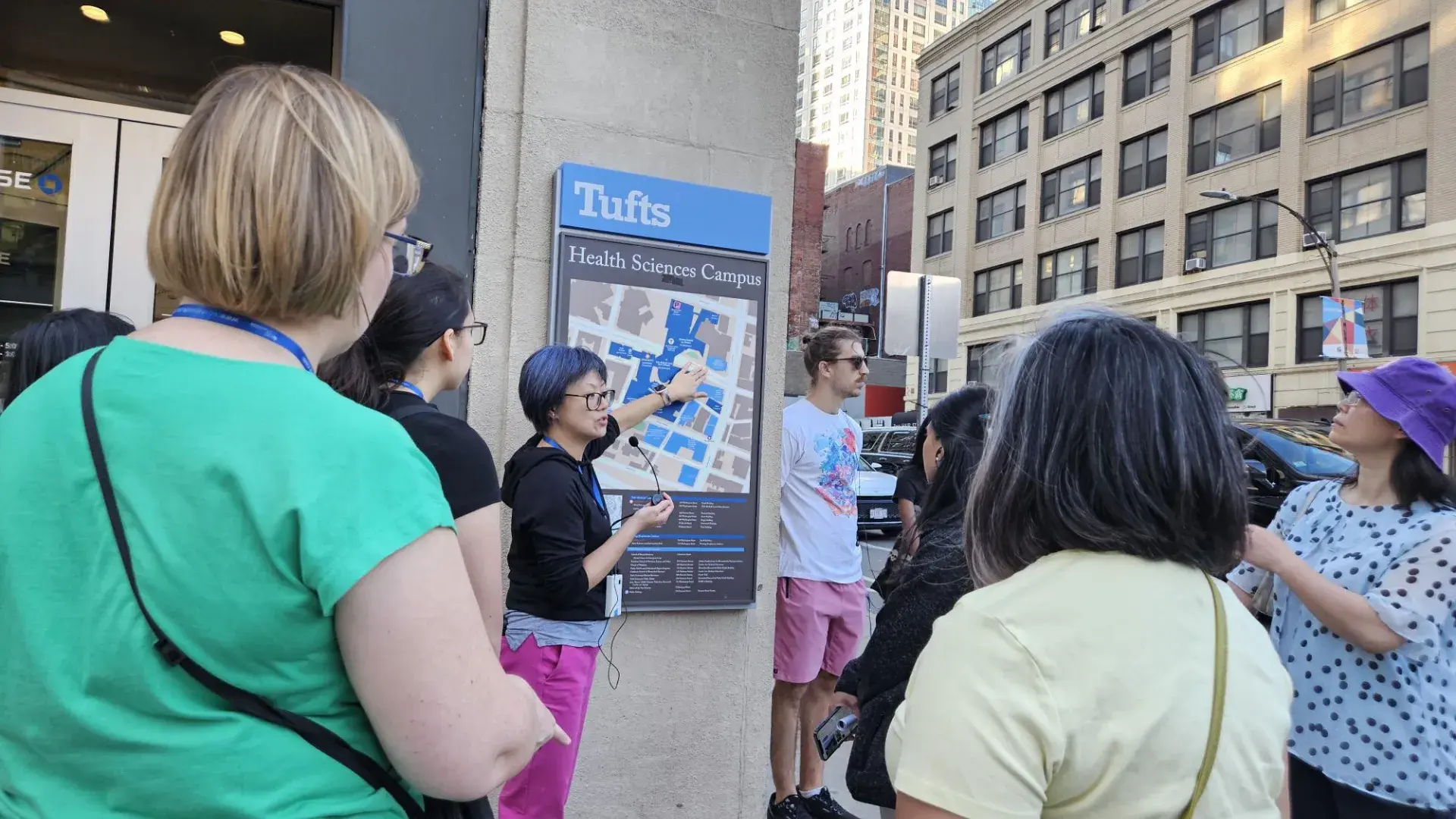 A group of people listen to a woman pointing to a map on a building under a sign that says Tufts