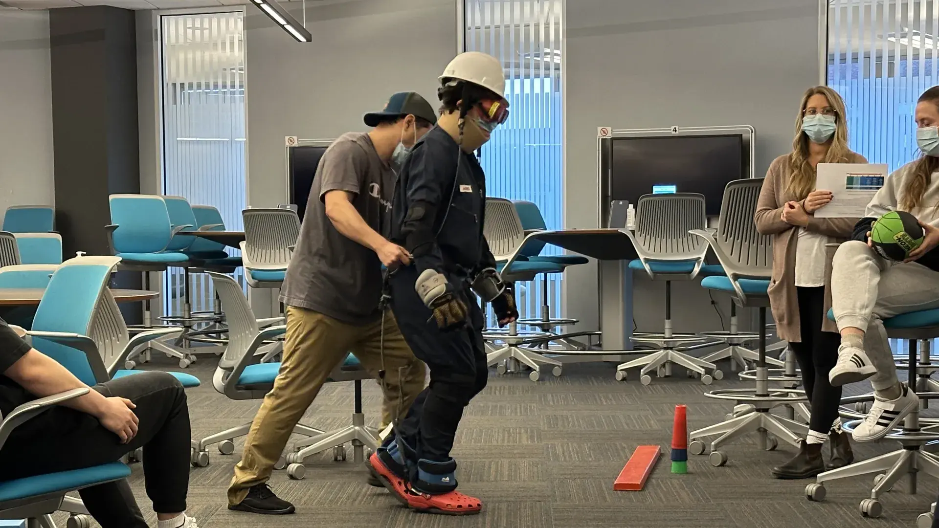 A person wearing a hard hat, goggles and gloves is assisted by another person while people watch