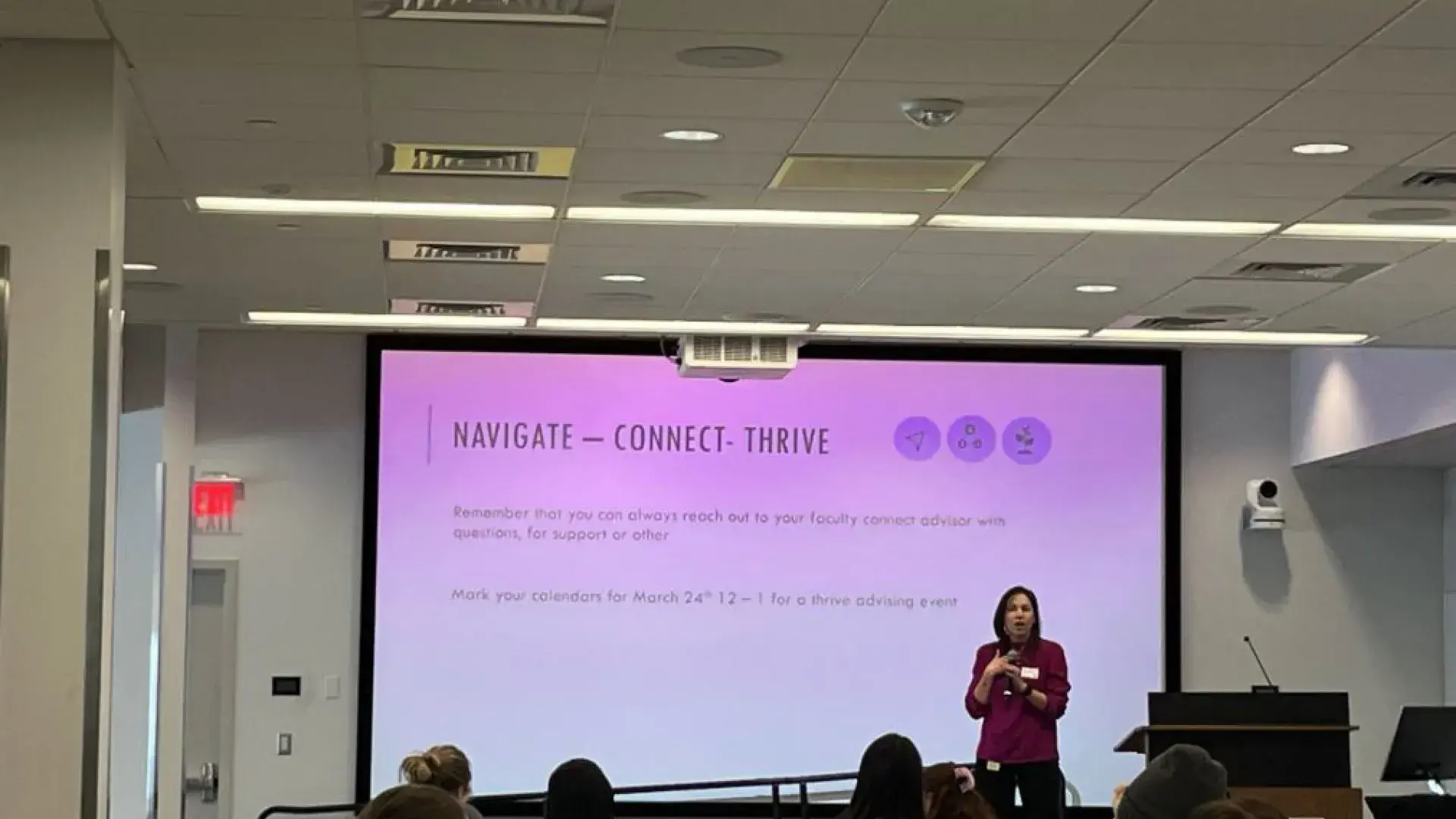 A woman stands on stage in front of a screen while people at tables sit in front of her. 