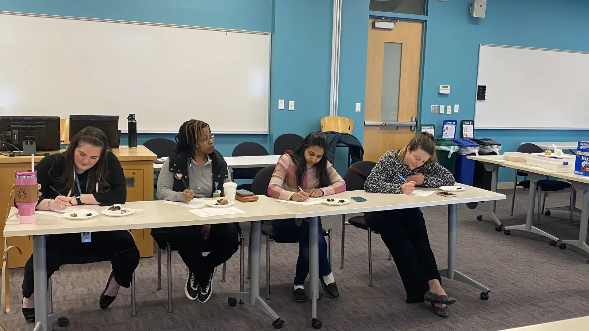 Four women write on paper in front of them while a fifth looks on