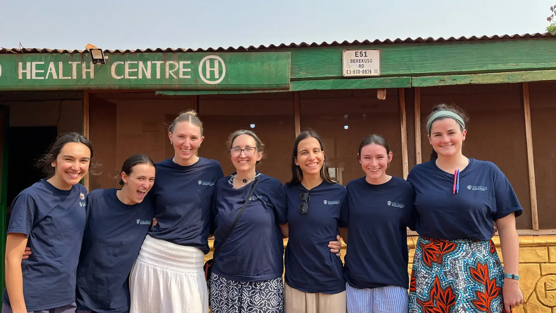 Seven women stand next to each other posing for a photo in front of a health center