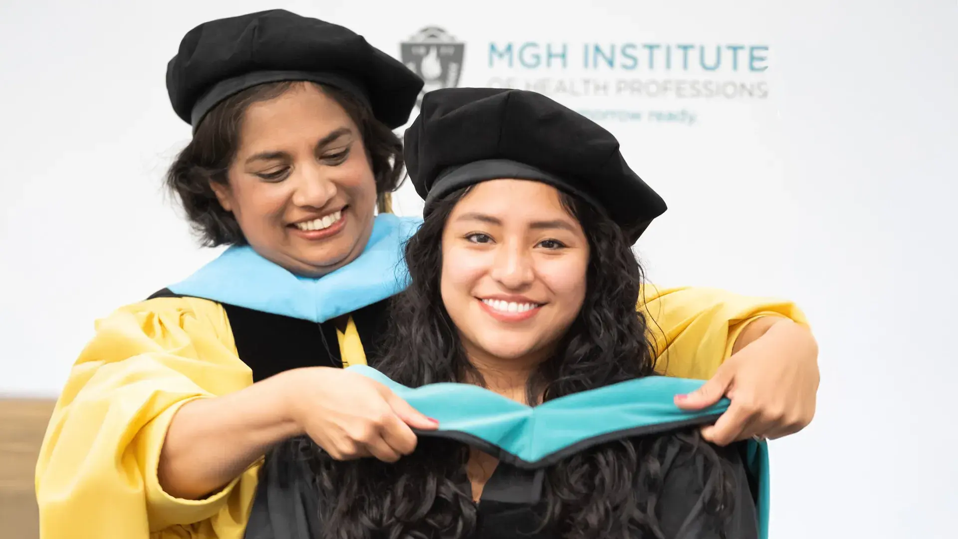woman puts a sash over another woman's head both wear graduation caps and gowns