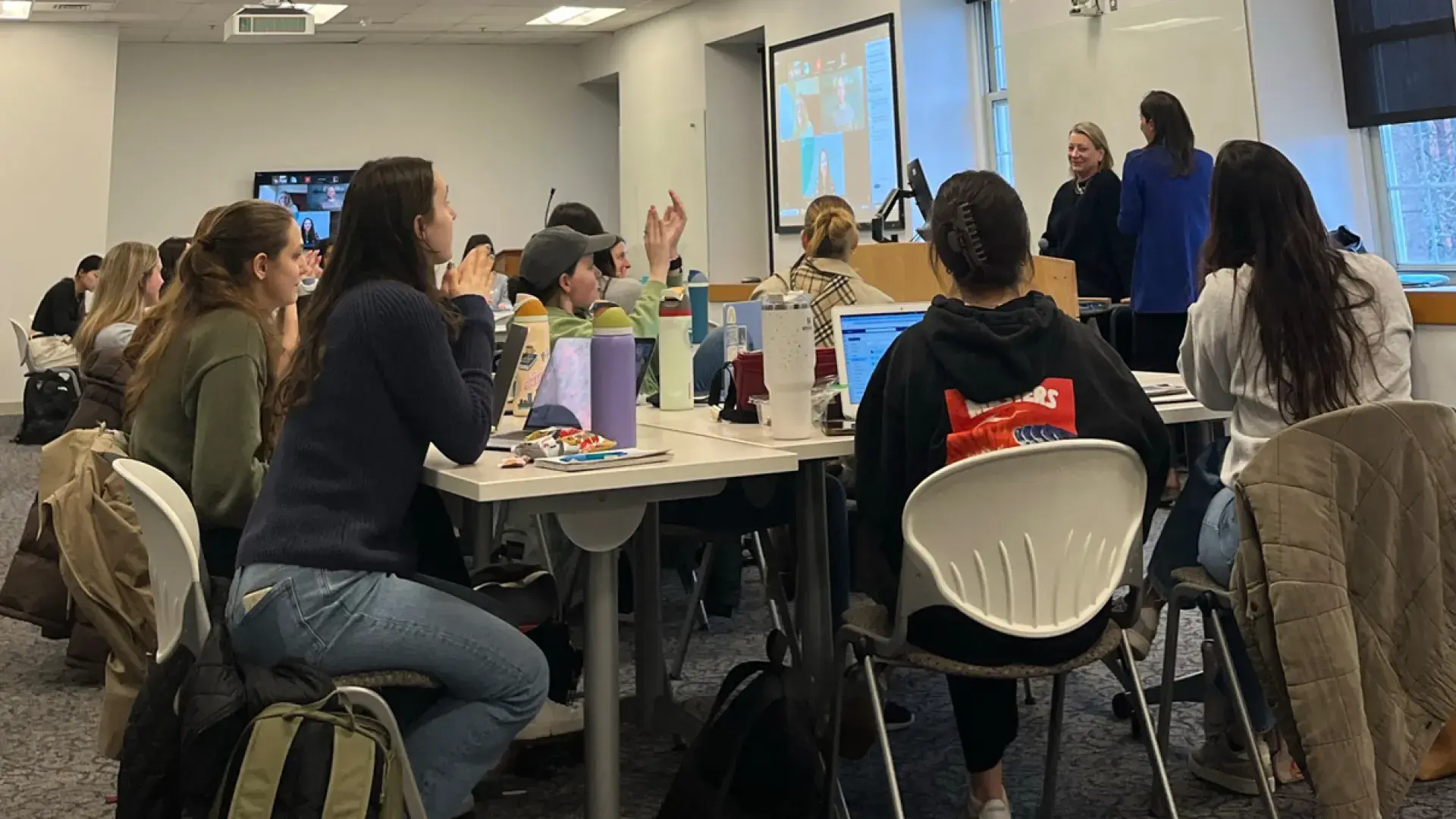 People sit at a table with computers in front of them with two people in the front of the room at a podium