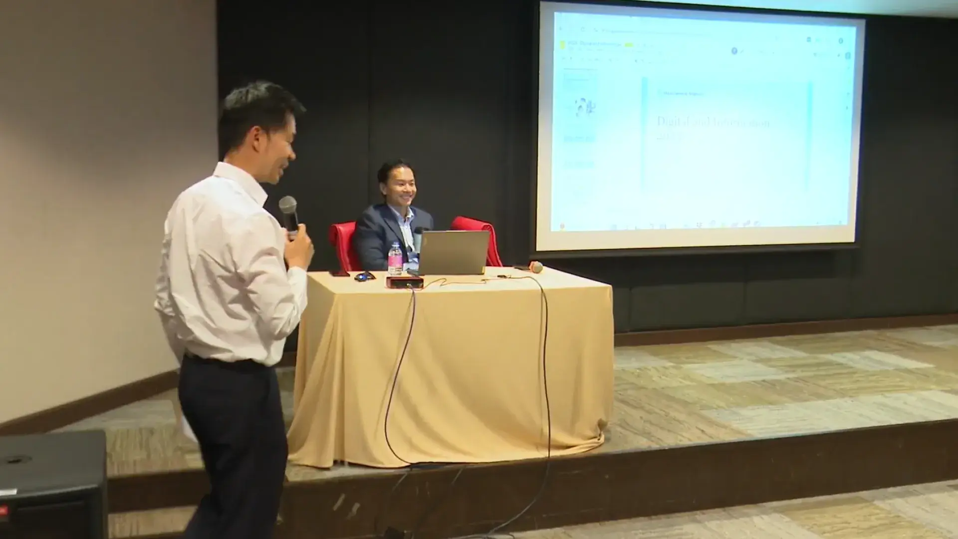 A man with a microphone stands in front of a table and screen