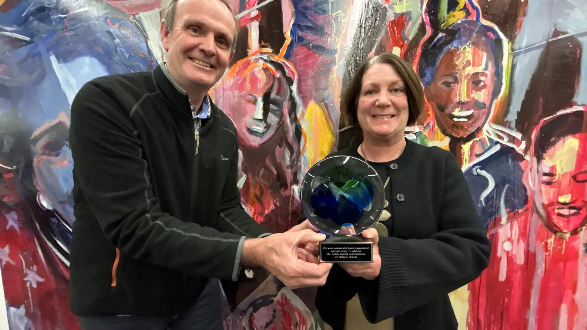 A man presents an award to a woman in front of a colorful mural on the wall