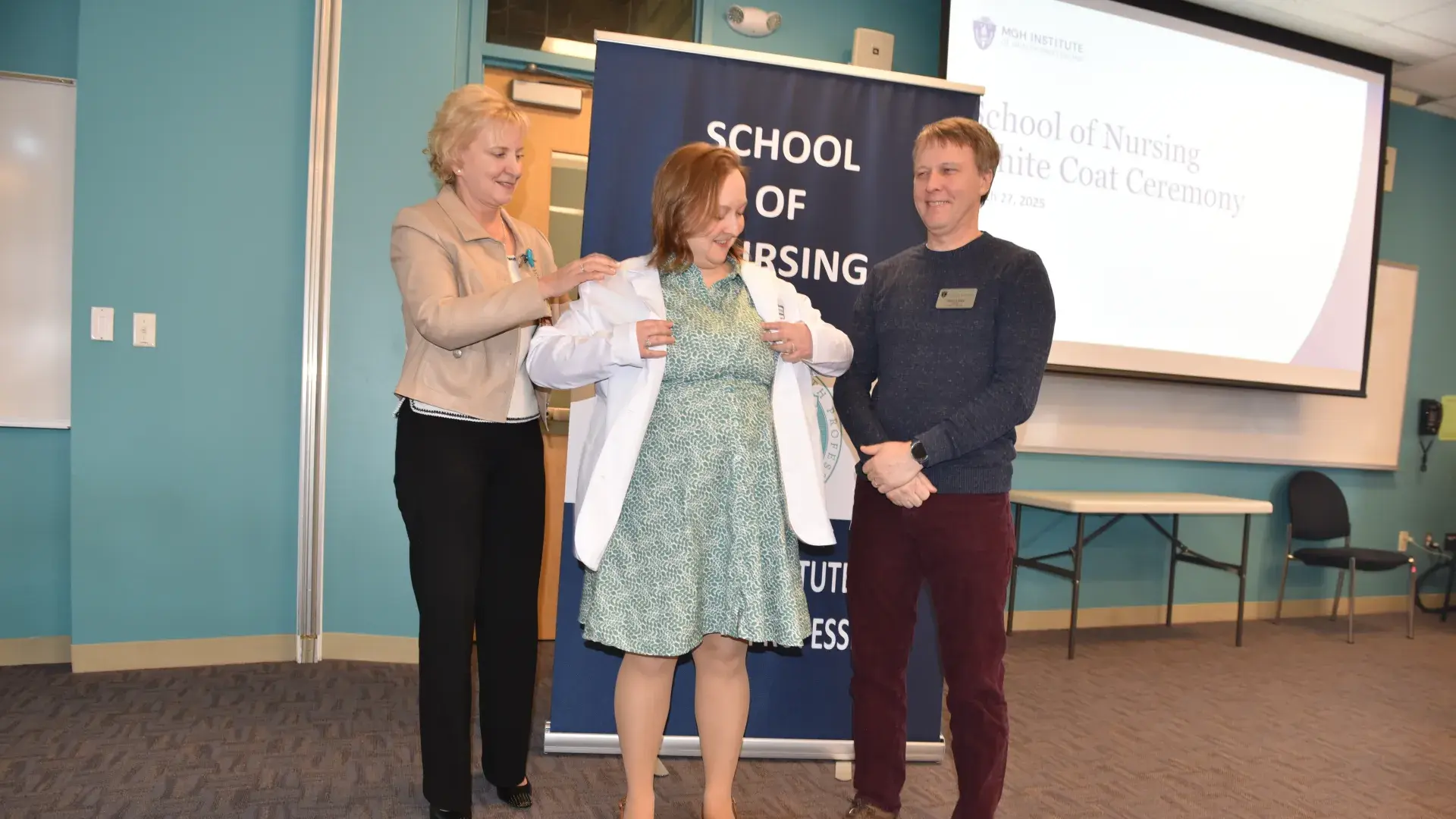 A woman helps another woman put on a white medical coat while a man observes