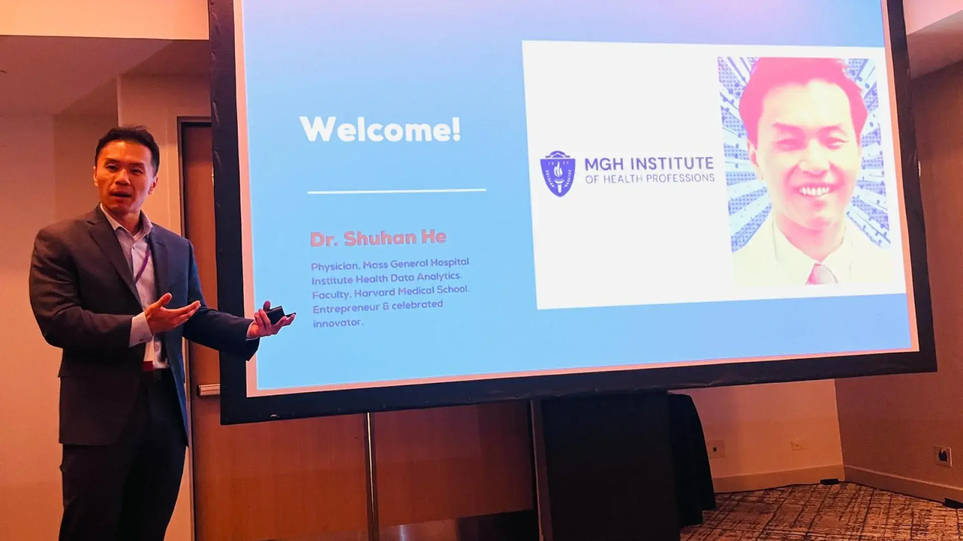 A man stands in front of a screen with his photo and the word Welcome on it. 