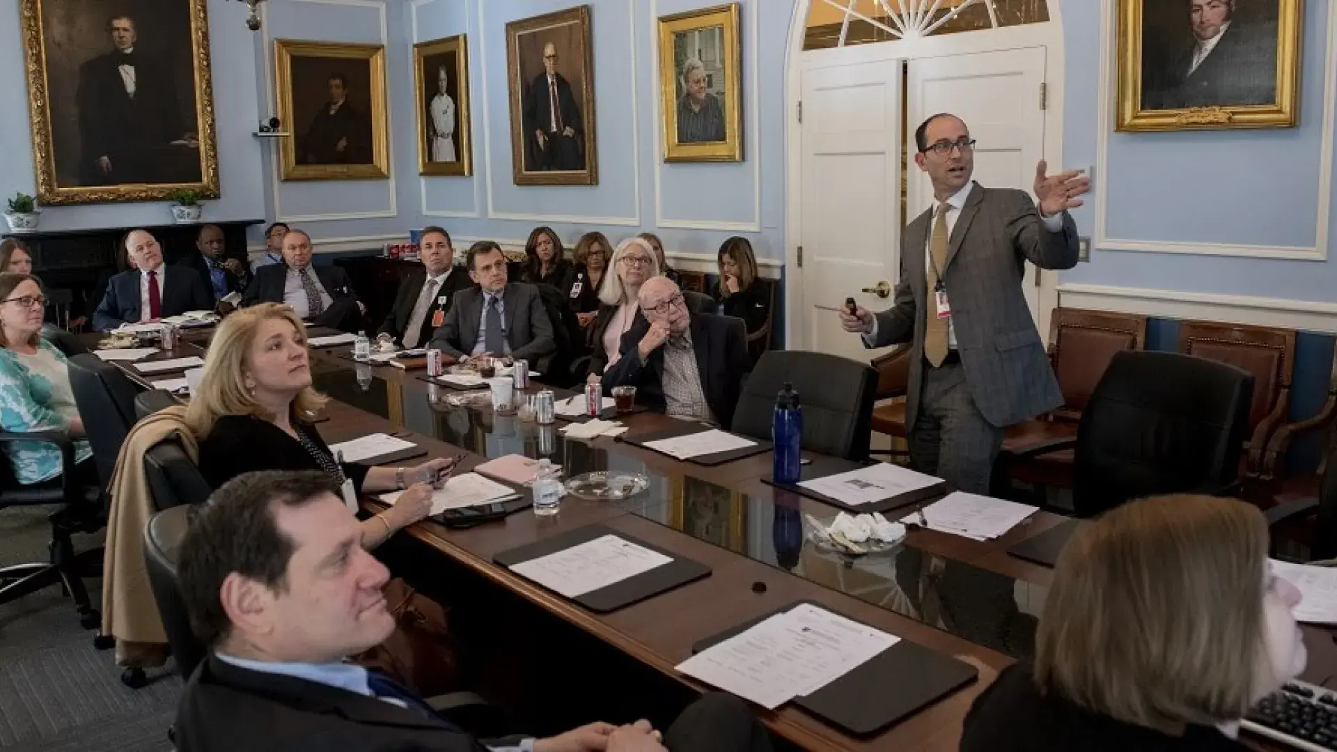 A man stands while others sit around a board table