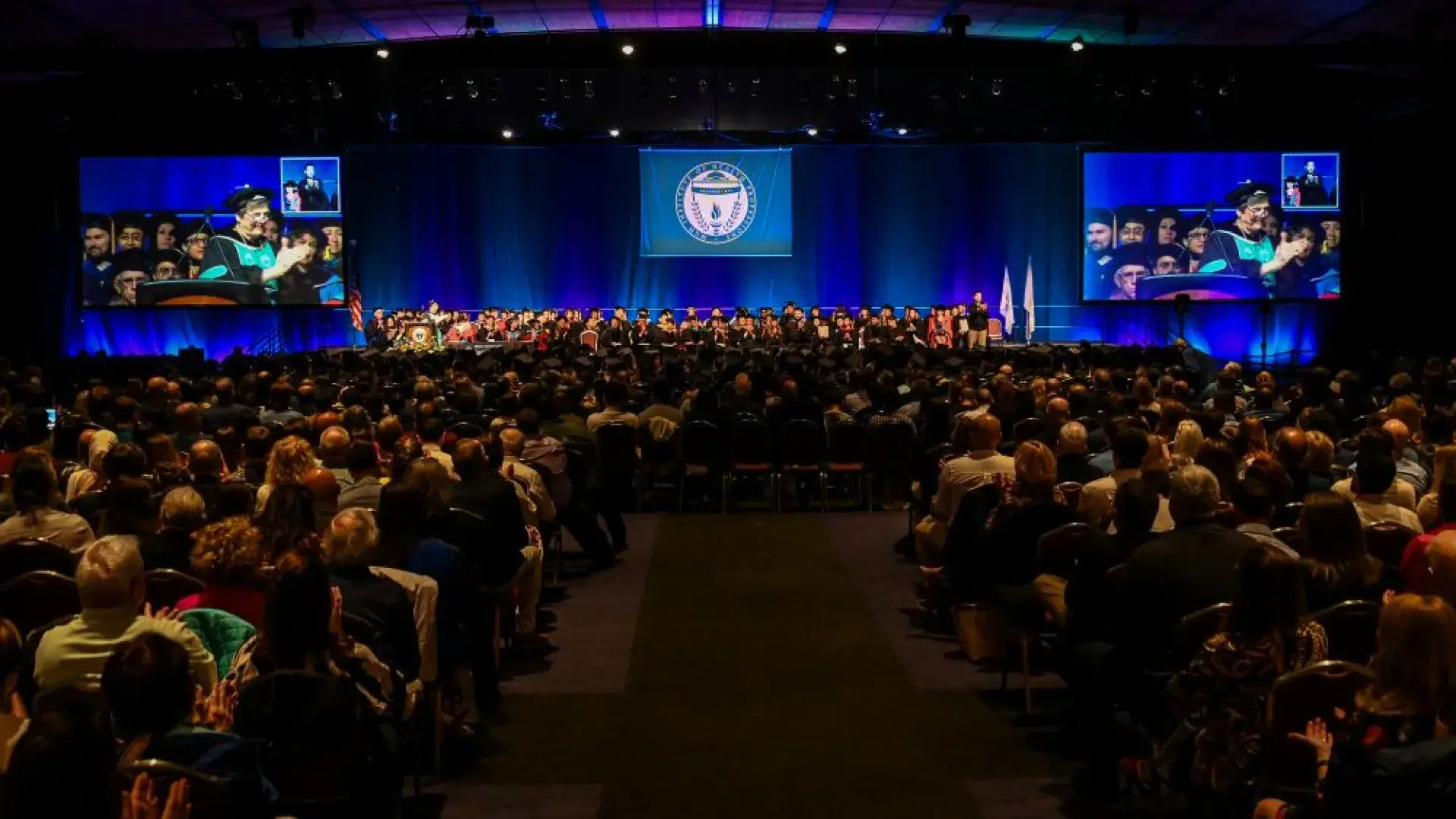 An audience sits in front of a stage while people wearing academic robes sits on the stage