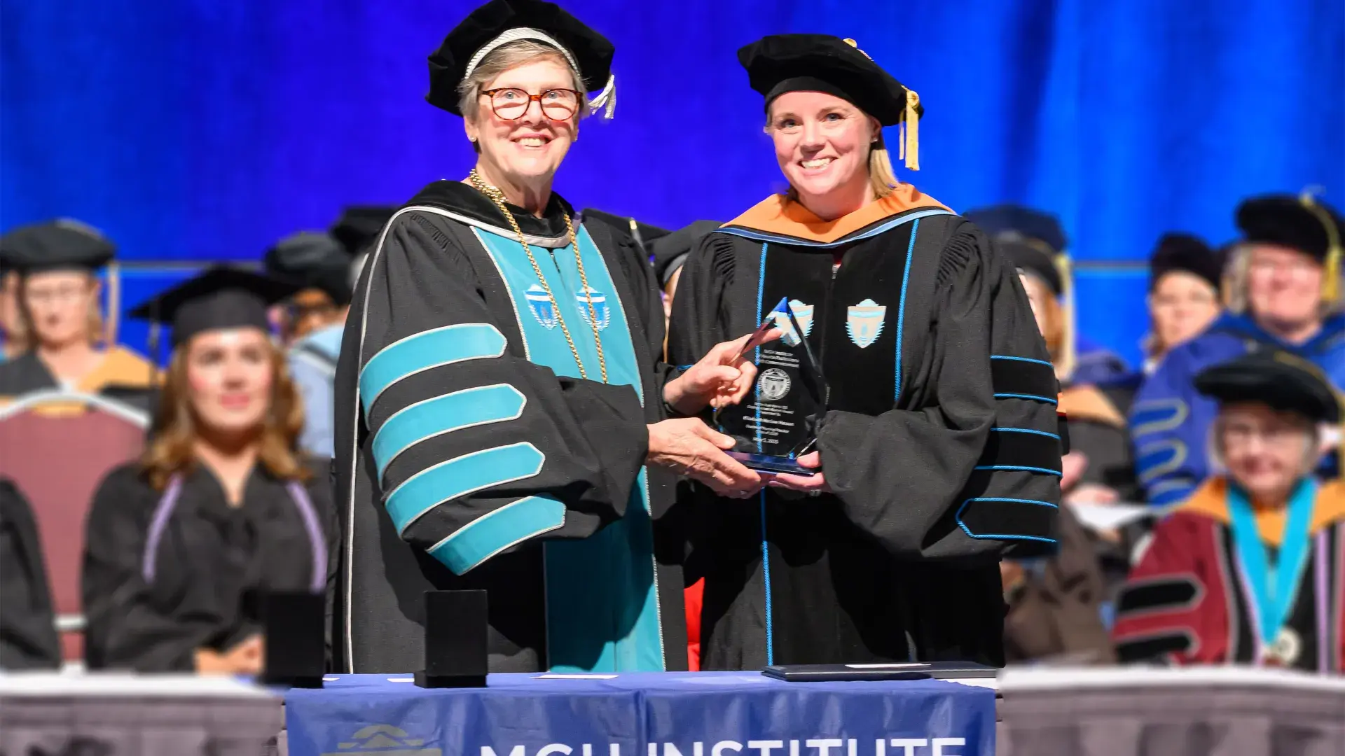 two women in graduation regalia hold a teardrop shaped crystal award