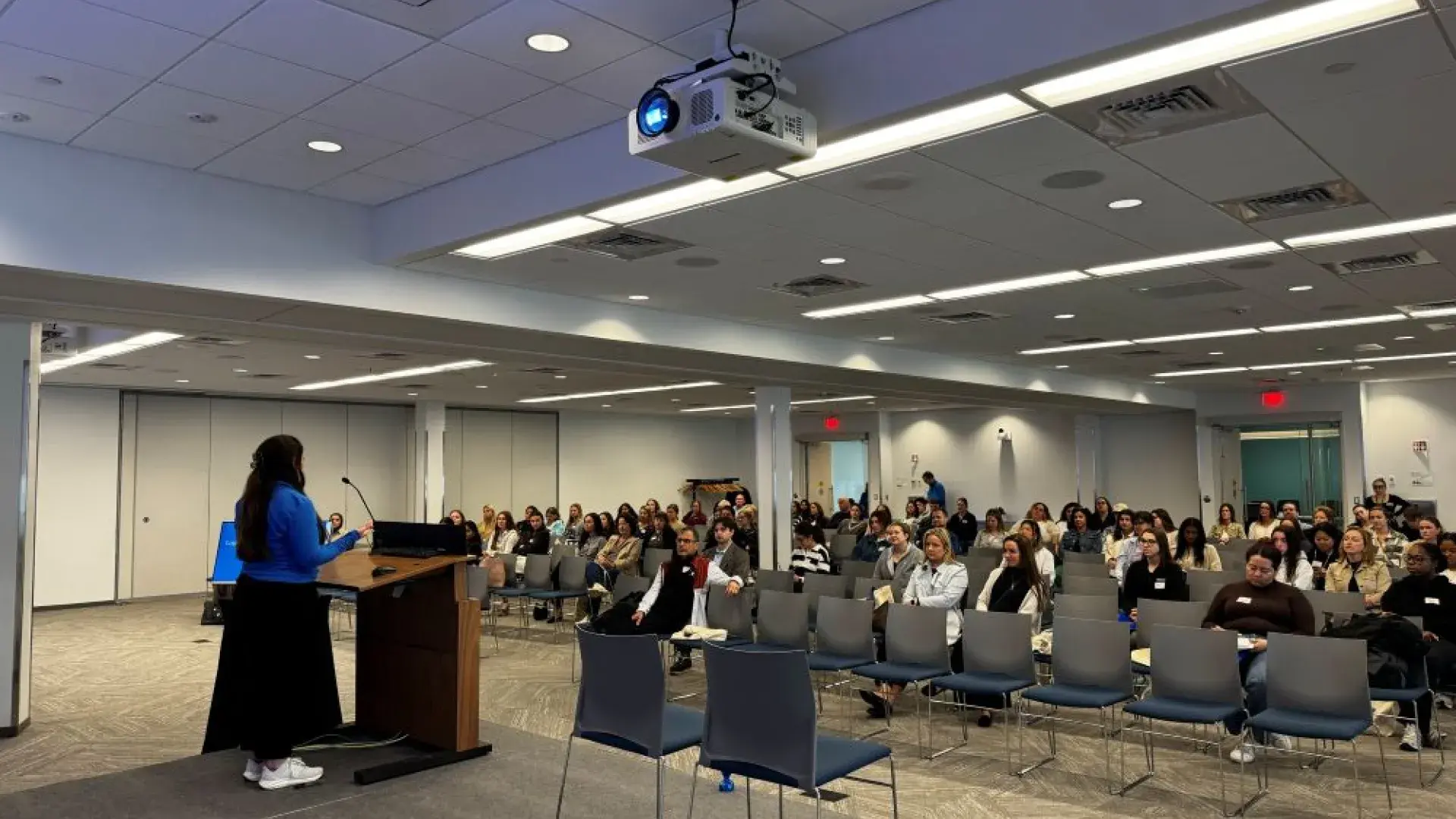 Woman standing on stage speaking to large audience in chairs
