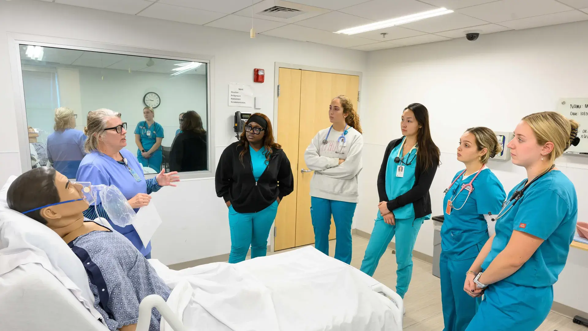 a woman in scrubs gestures to a manikin in a hospital bed while other people in scrubs look on