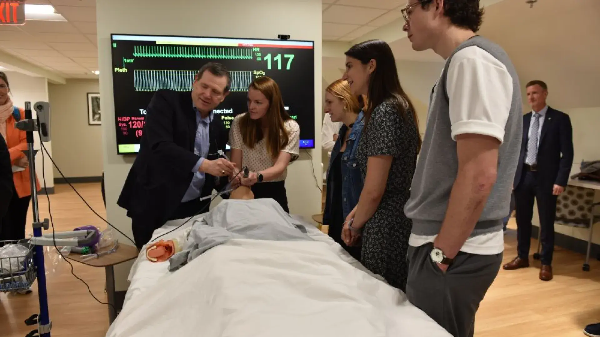 Two people look at a screen while working on a manikin in a hospital setting while three others look on
