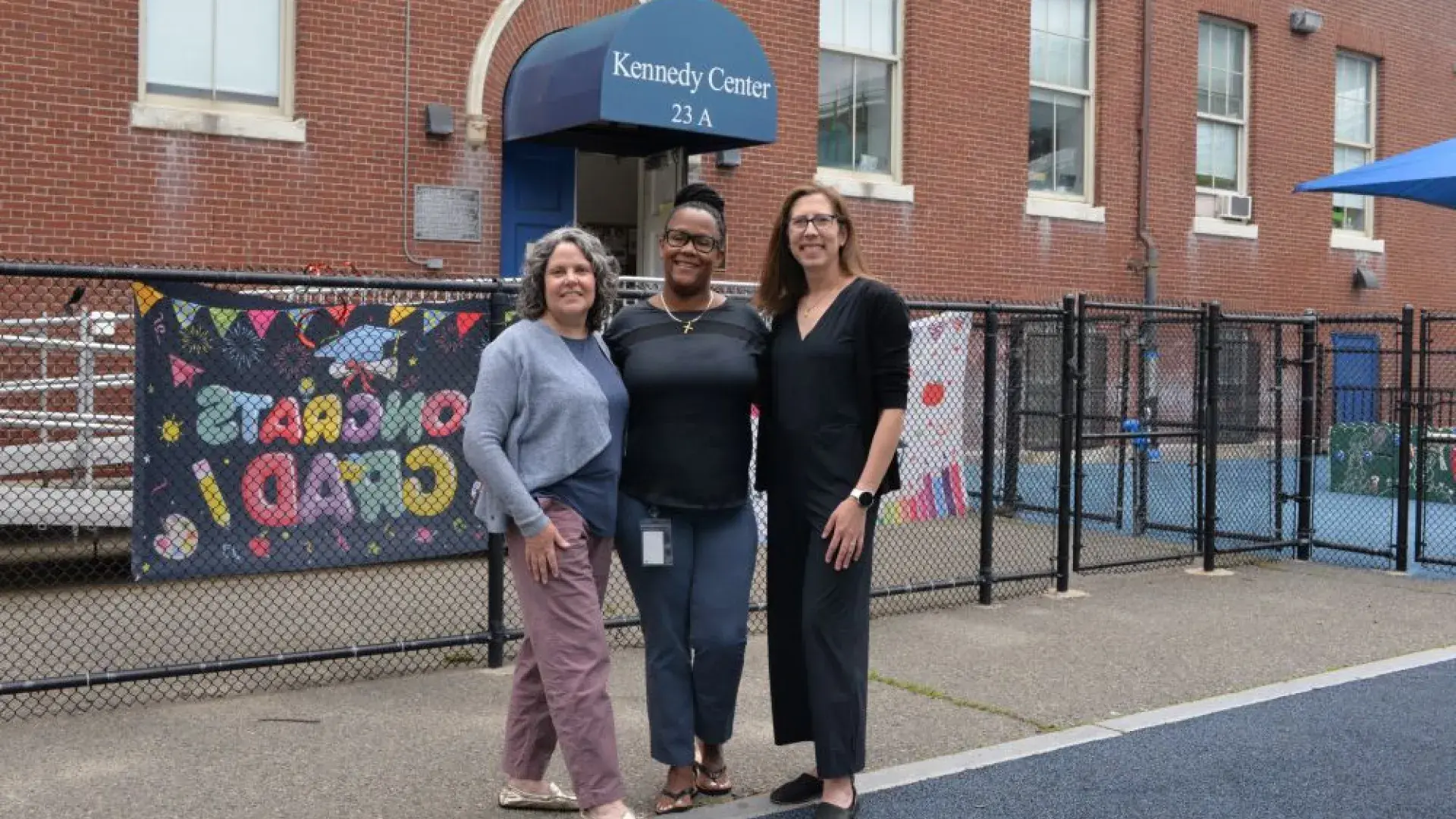 Three women pose outside a building that has a sign that reads Kennedy Center