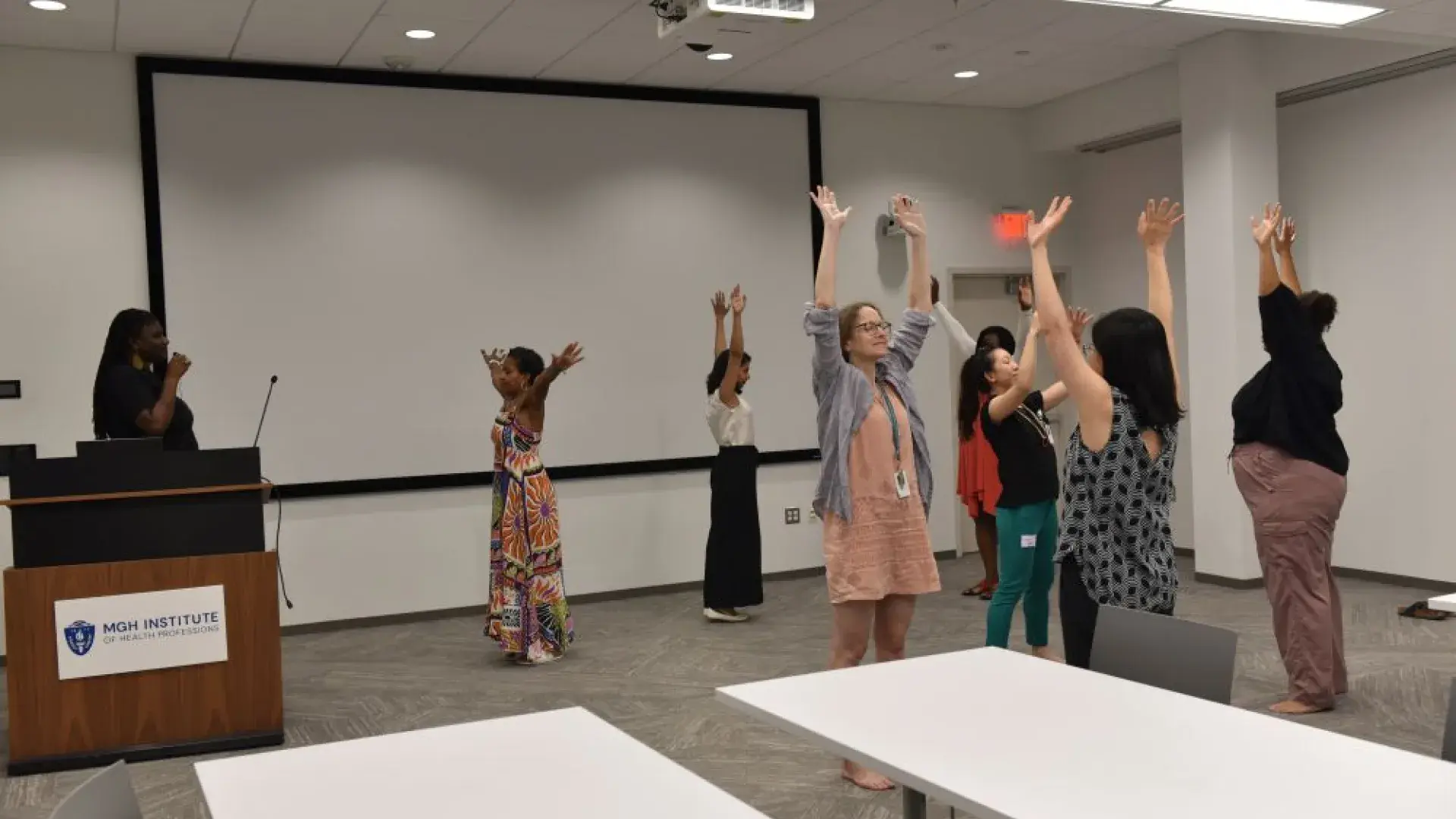 Several people stand facing different directions with their hands in the air while a woman with a microphone stands on the left