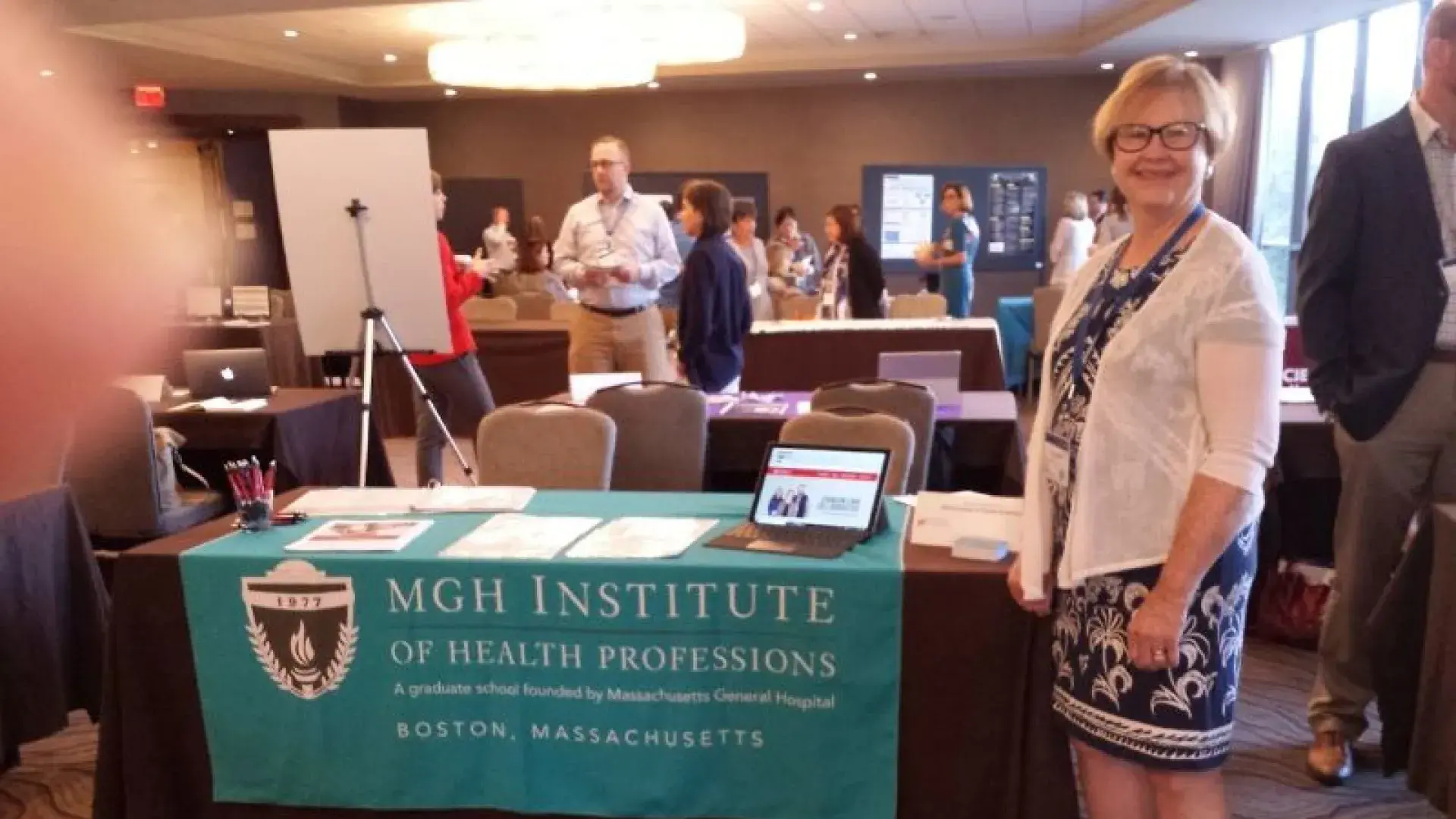 A woman stands in front of a table with a banner that says MGH Institute of Health Professions