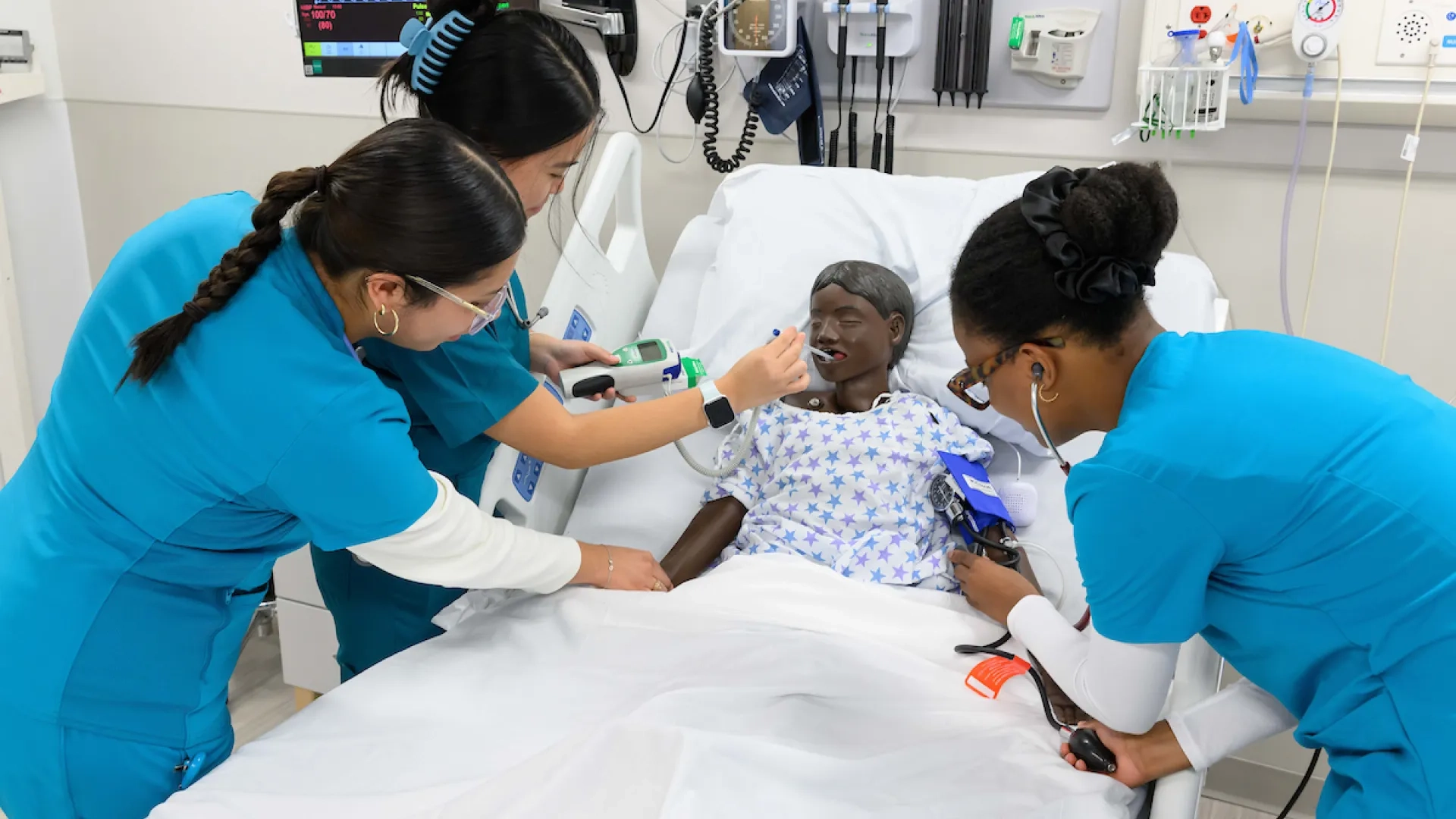 three women in blue scrubs surround a manikin child in a hospital bed