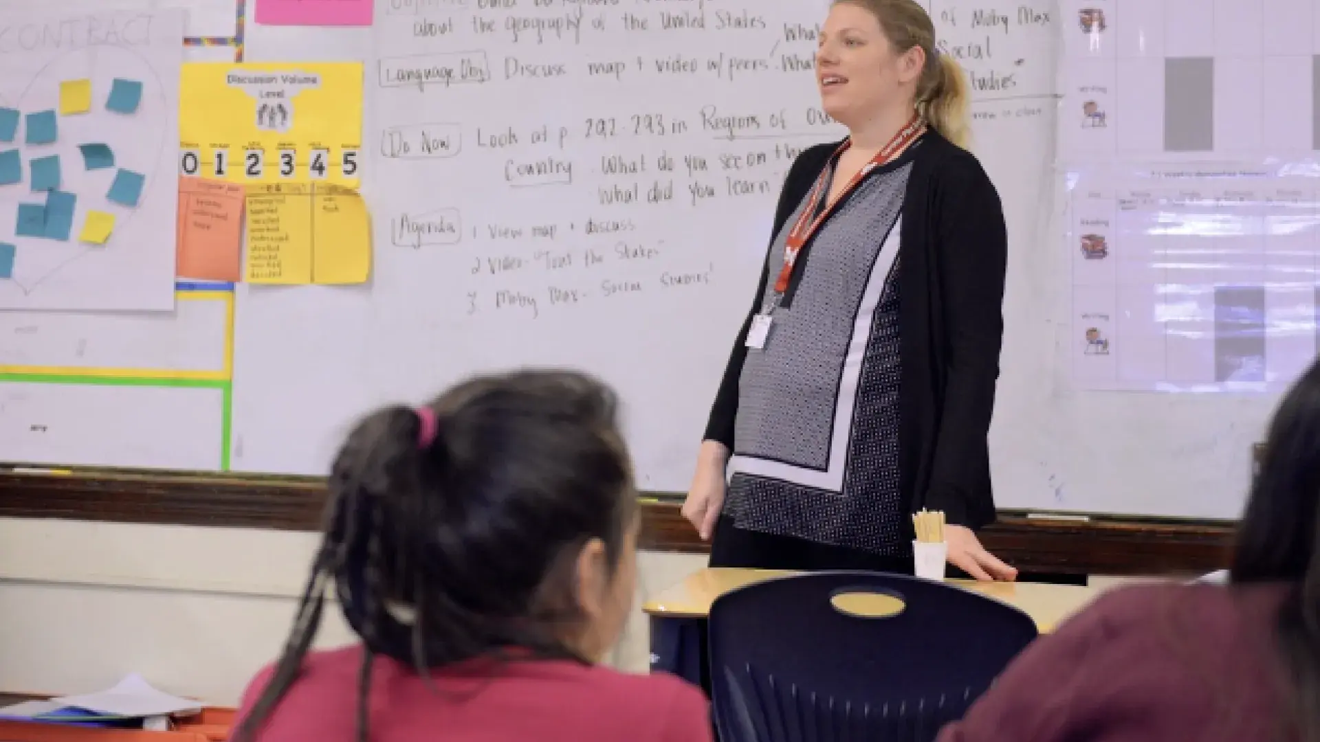 a woman stands in front of a classroom white board with the back of student heads in the foreground