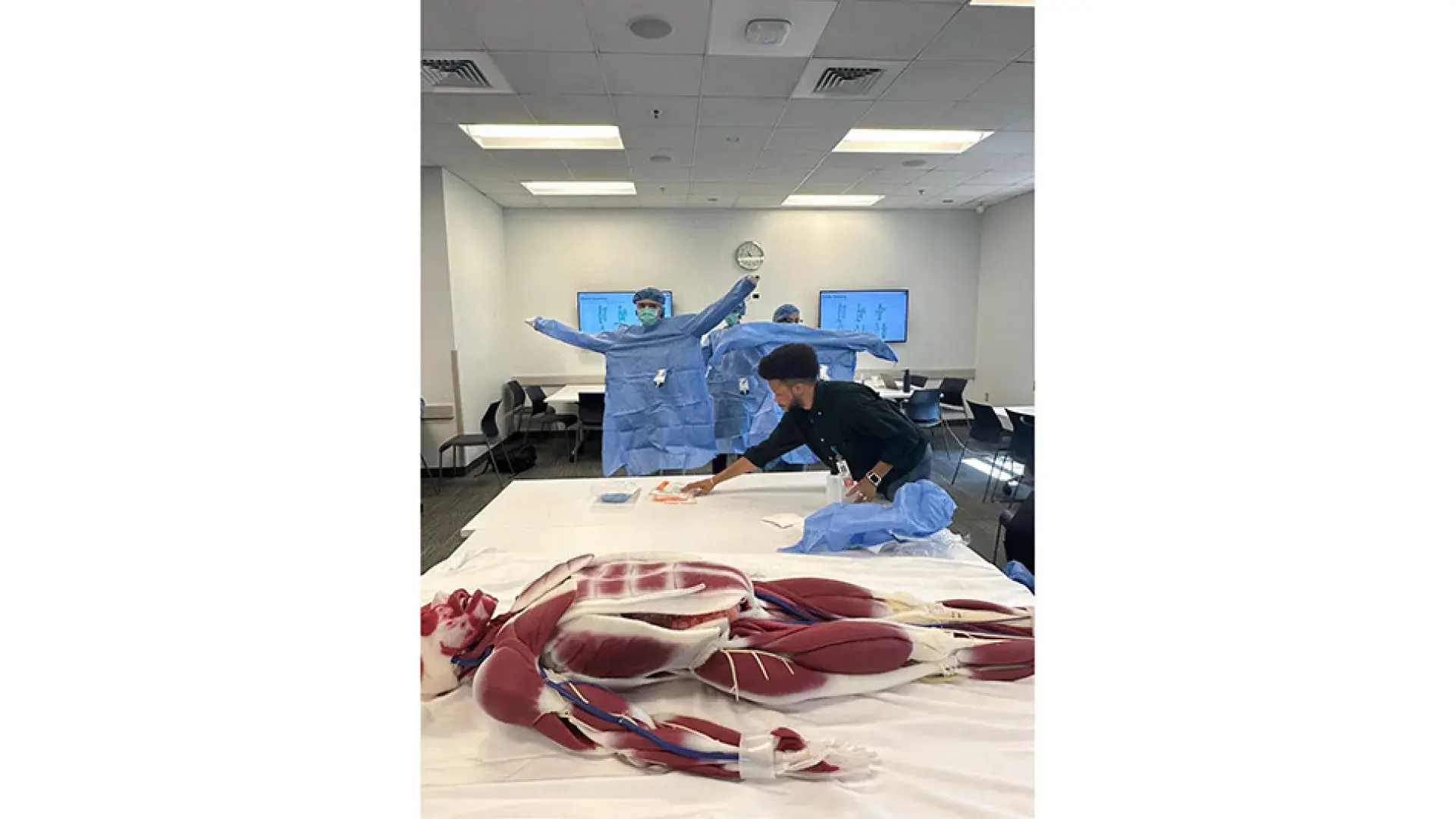An anatomical manikin showing a body's muscles lies on a table while students don blue medial gowns