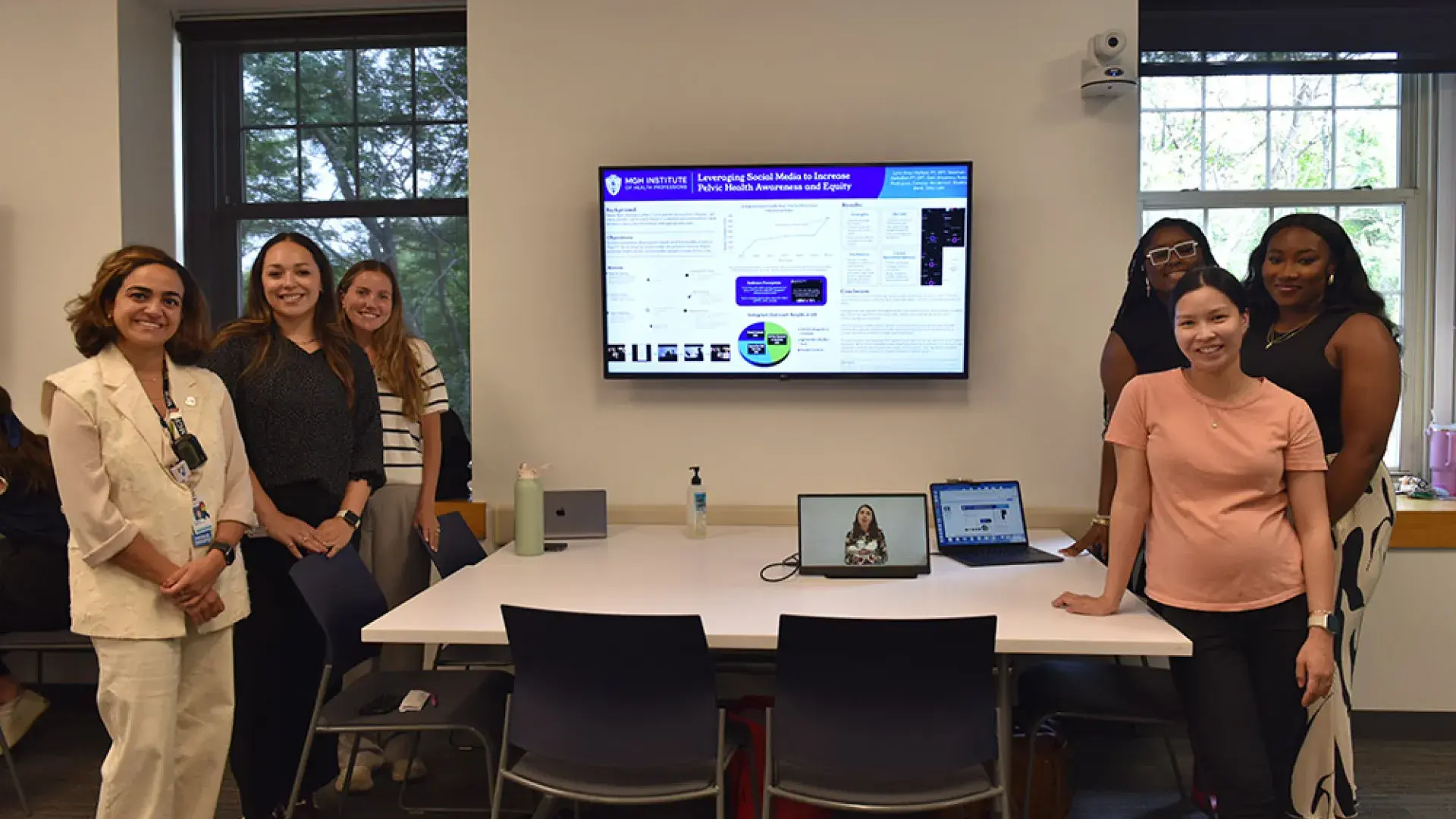 Six people stand around a table with a tv screen behind them showing a presentation