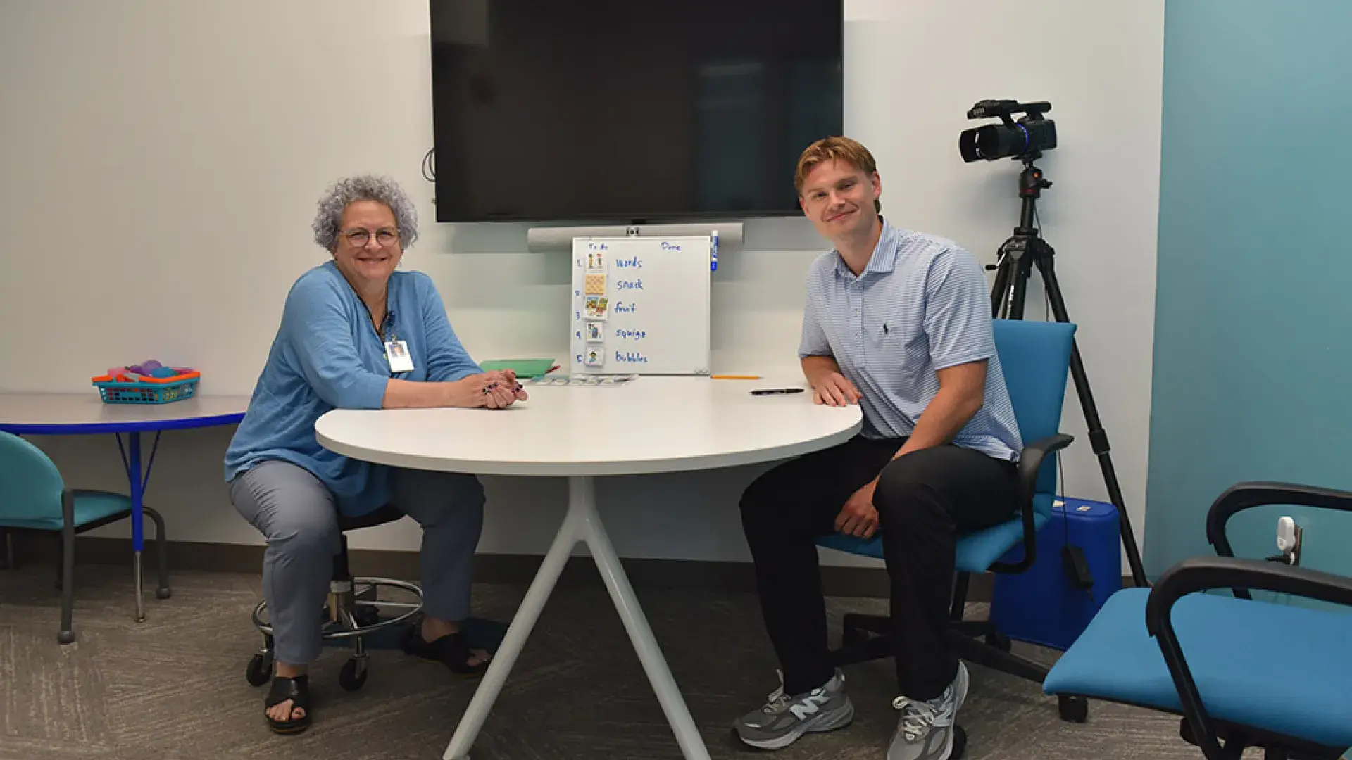 Two people sit at a table with a tv screen behind them 