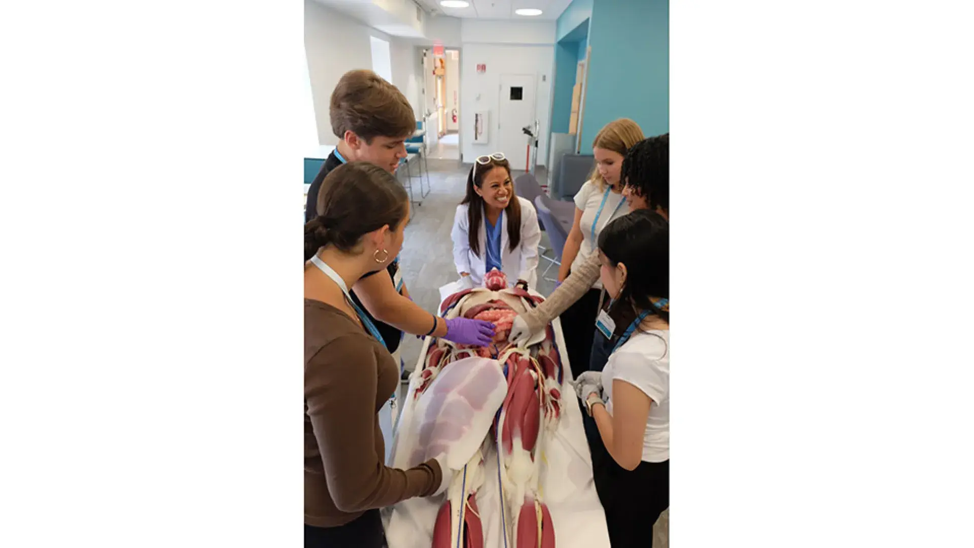 Students look over a medical manikin on a table while a teacher talks to them from the head of the table