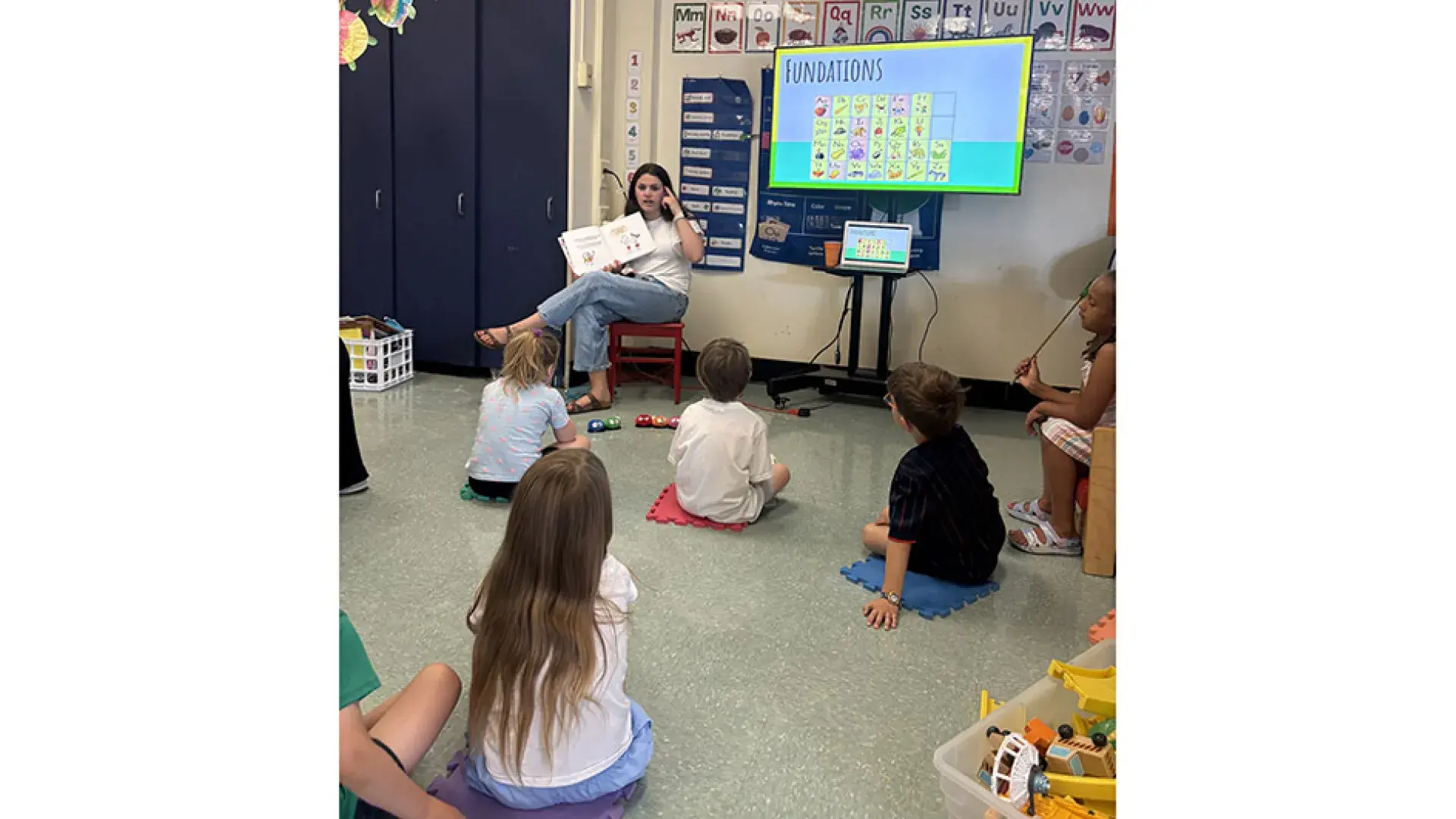 A woman sits in a chair with children sitting on the floor in a classroom
