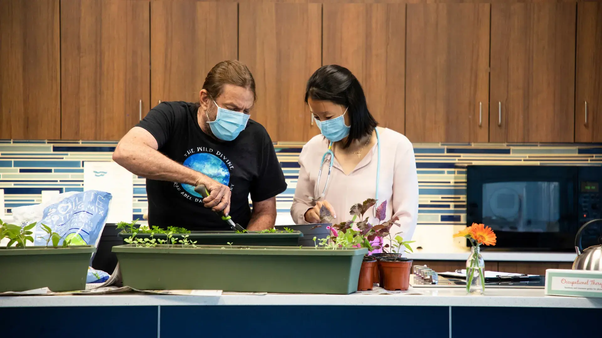 woman wearing mask and stethoscope helps middle aged man plant flowers in a pot