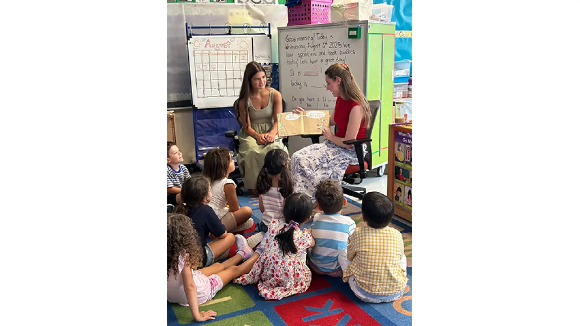 Two women read aloud to children sitting on the floor of a classroom