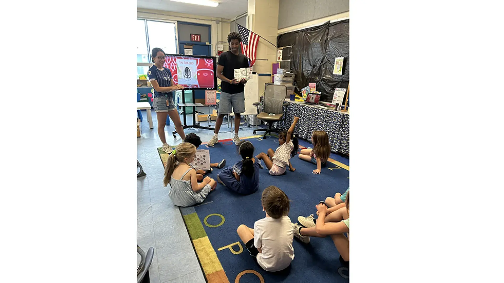 A person stands with a book in their hands in front of kids sitting on classroom floor while another adult looks on