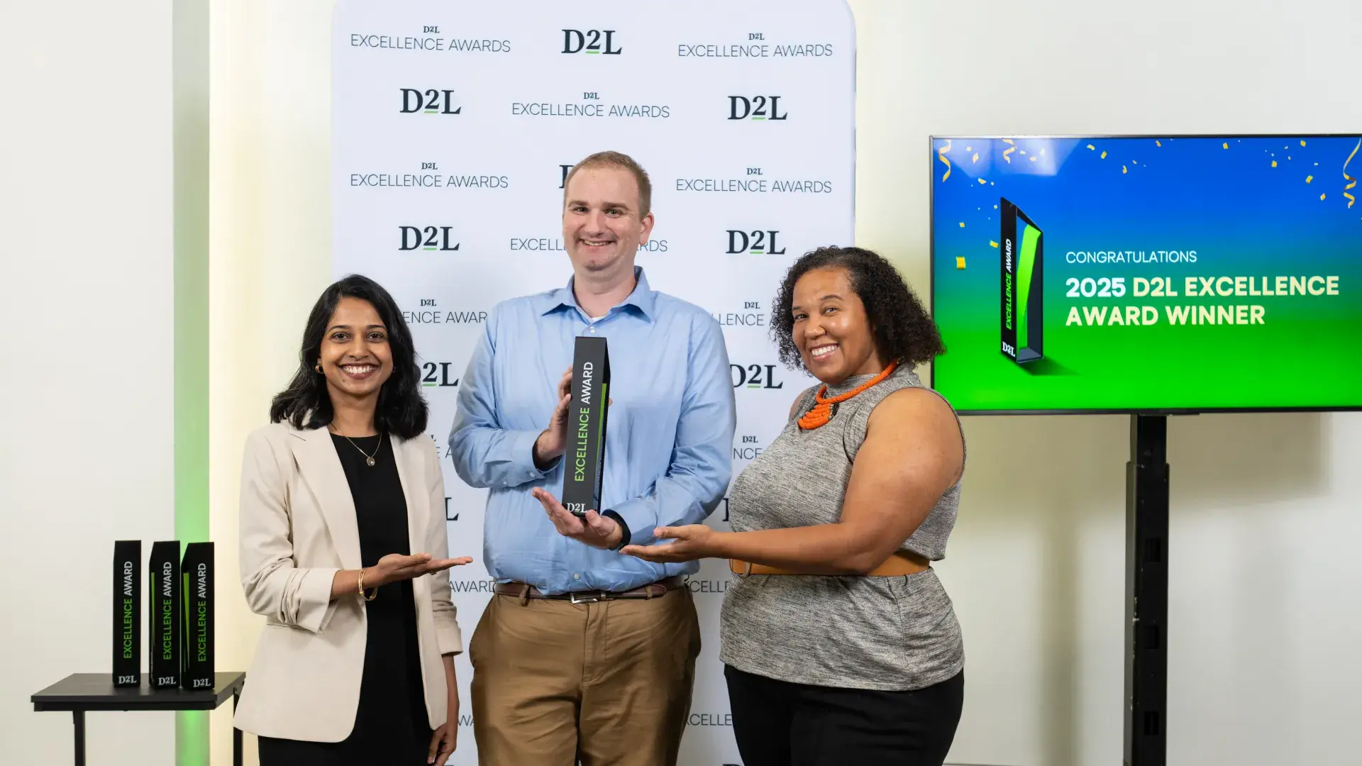 Three people stand in front of a banner with the man in the center holding a trophy