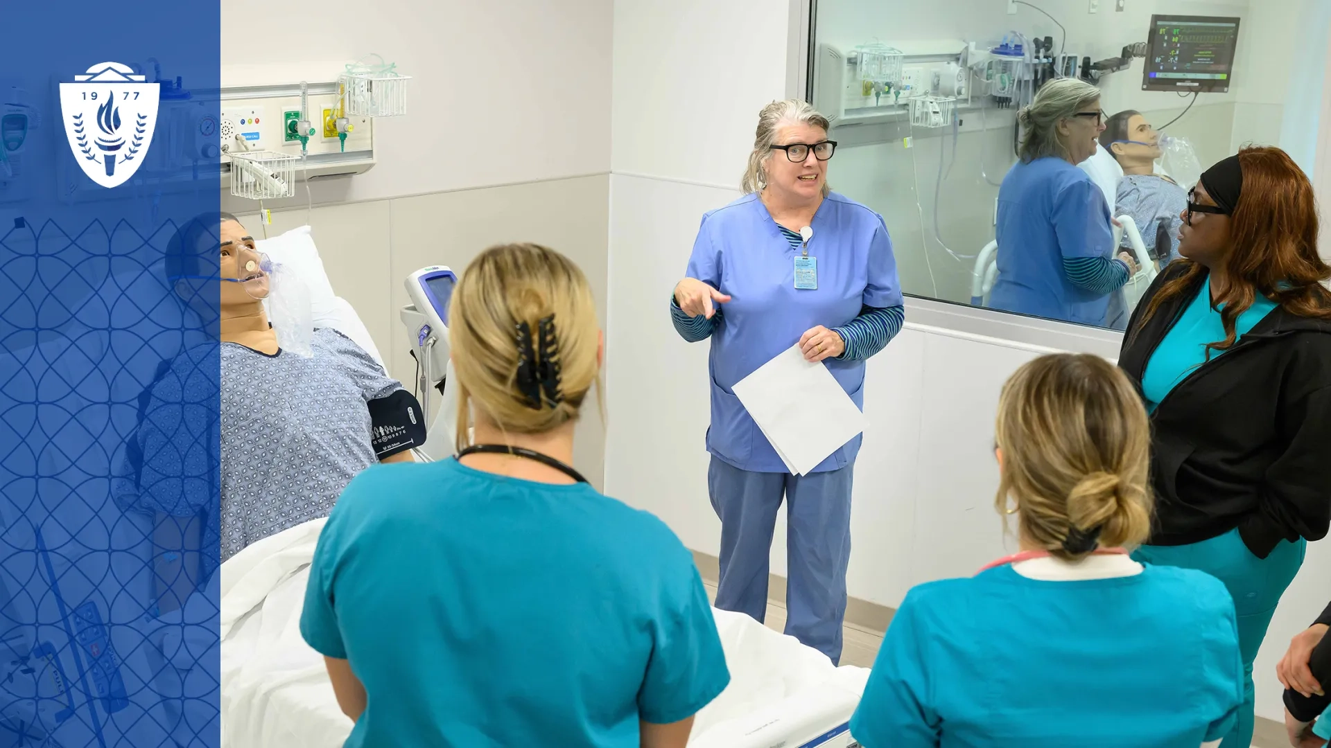 Professor and students wearing scrubs standing around a simulation hospital bed with a mannequin laying in the bed