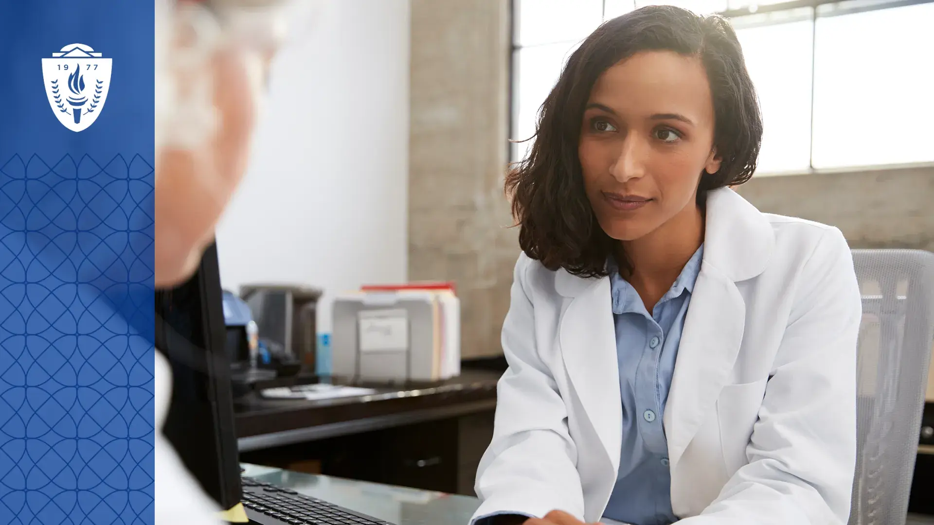 Woman wearing blue shirt and white lab coat sitting at desk accross from another person