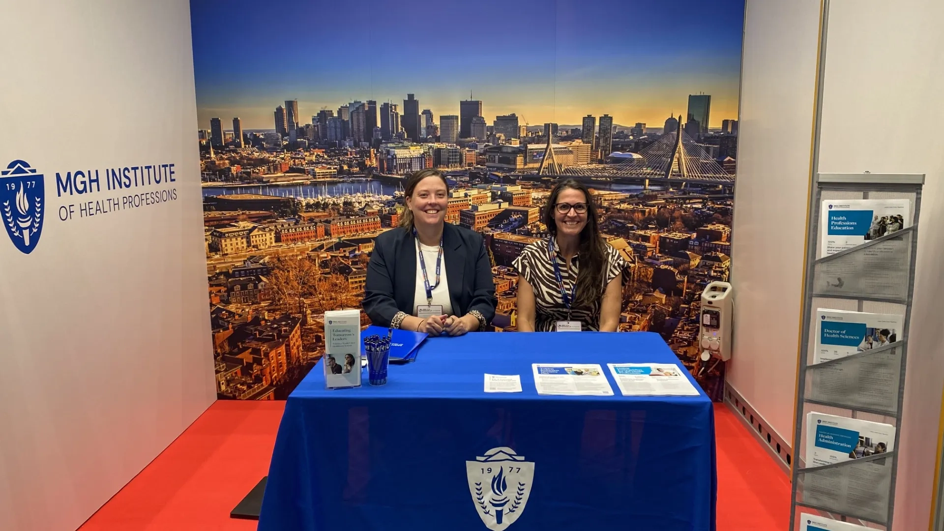two women sitting at MGH Institute booth with a background of Boston