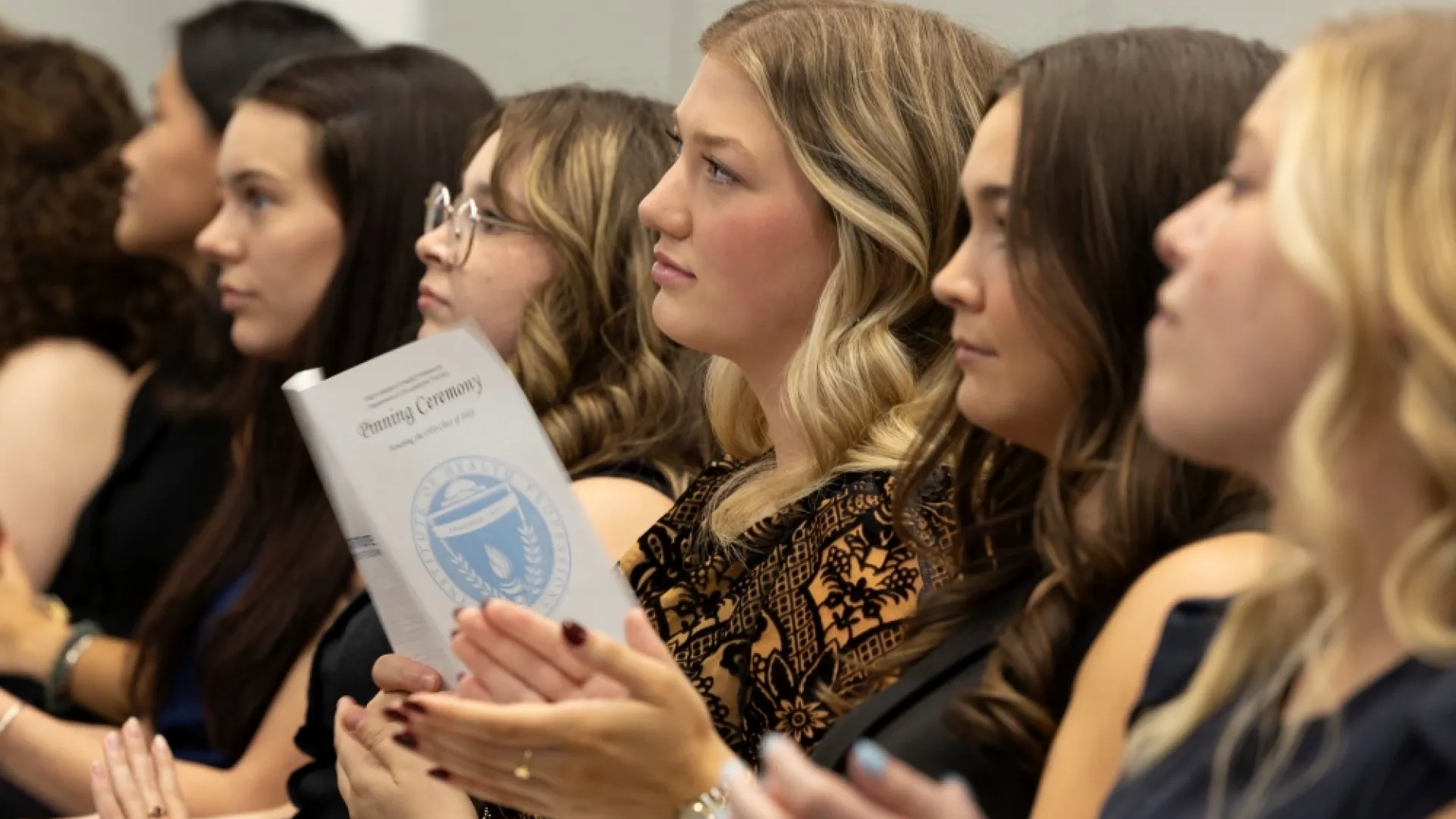 People sit in a row watching while one holds a piece of paper that says Pinning Ceremony