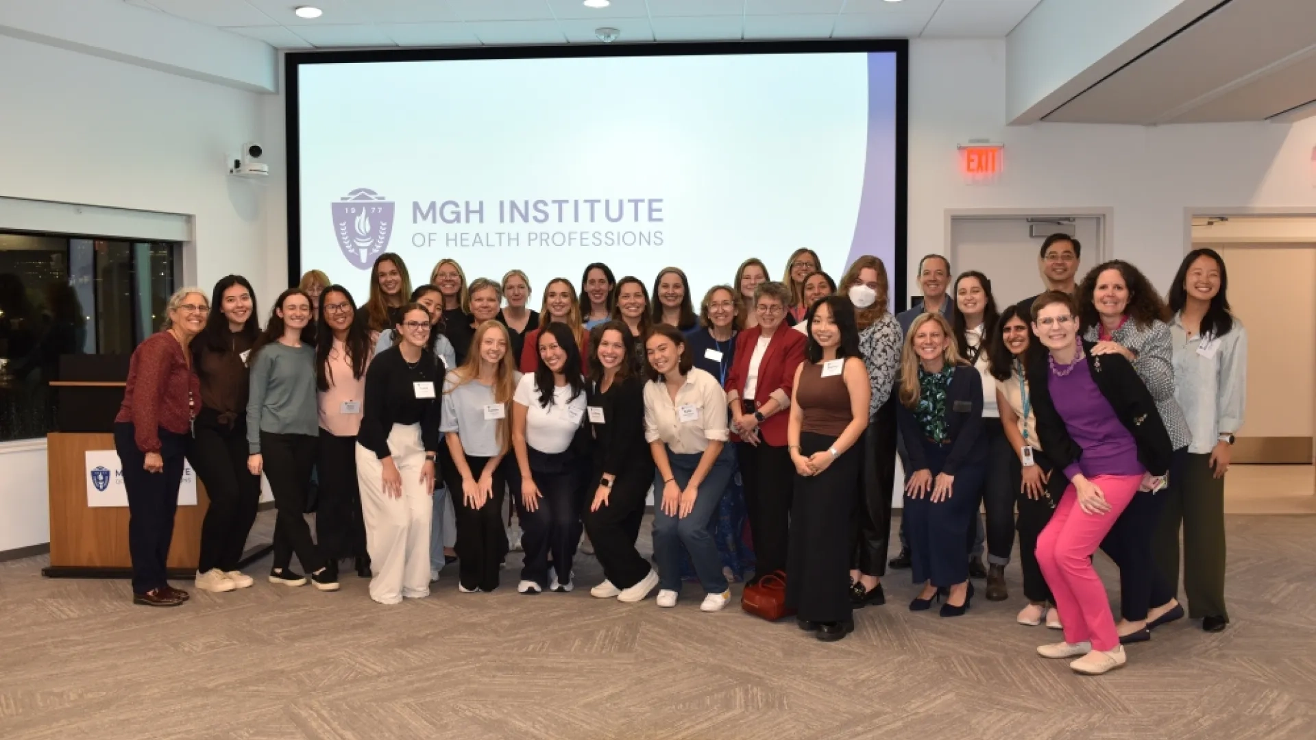 A large group of people stand together for a photograph in front of a screen that says MGH Institute of Health Professions