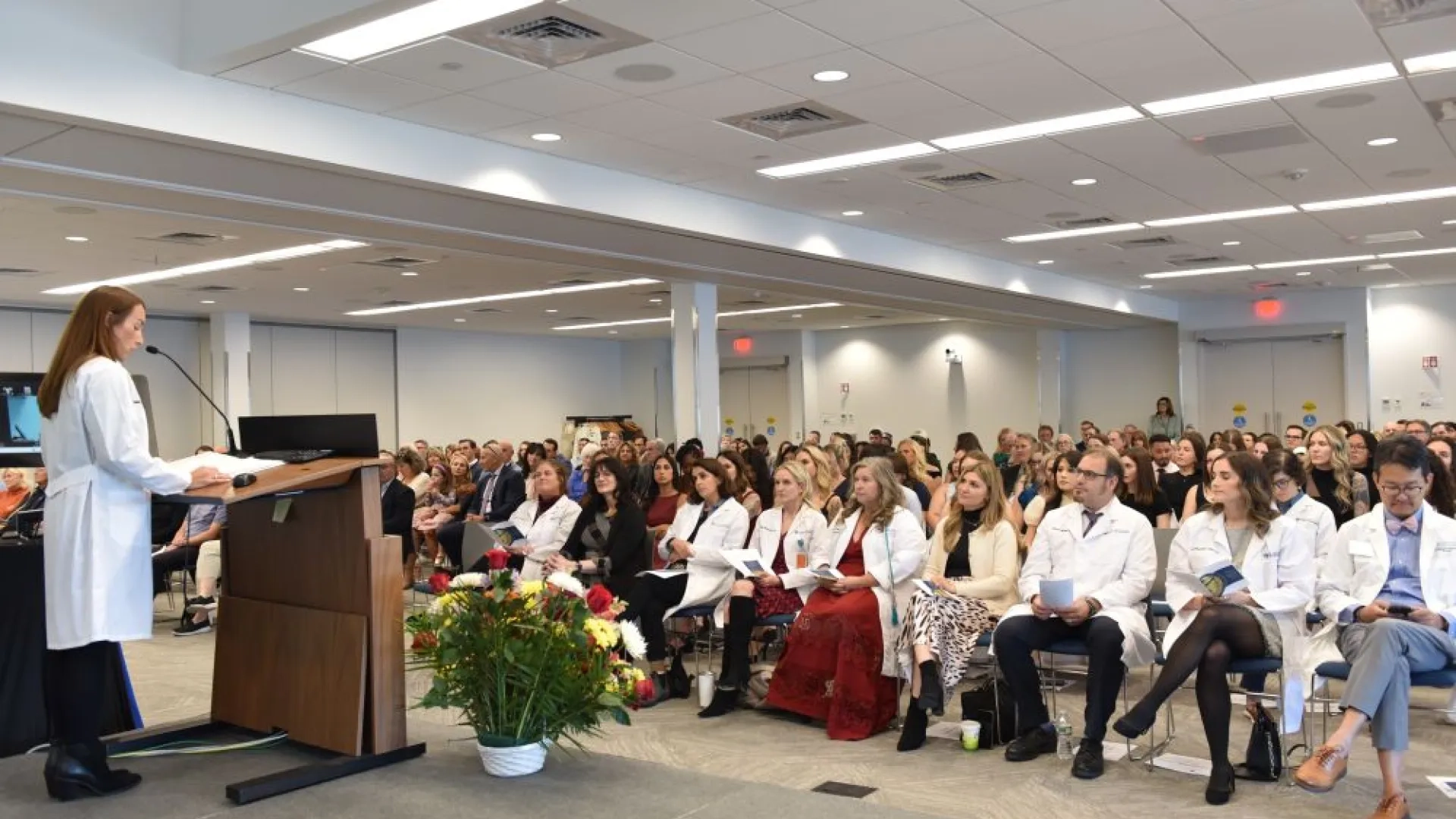 Woman standing at a podium addressing tens of people