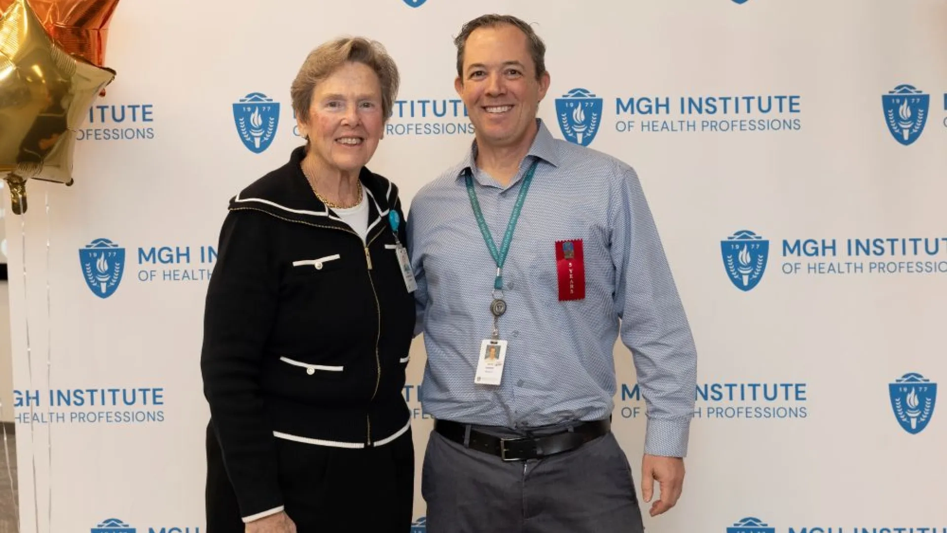 Two people pose in front of an MGH Institute backdrop