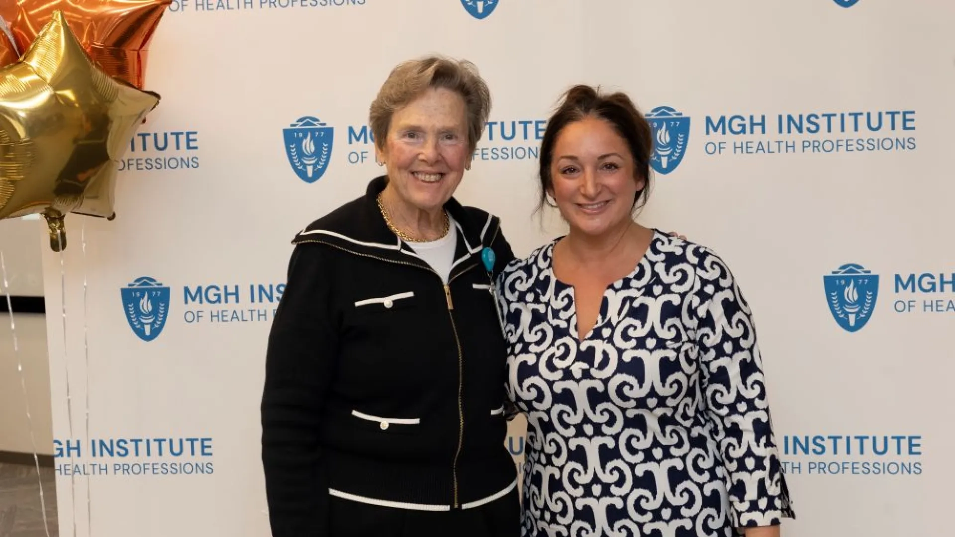 Two people pose in front of an MGH Institute backdrop
