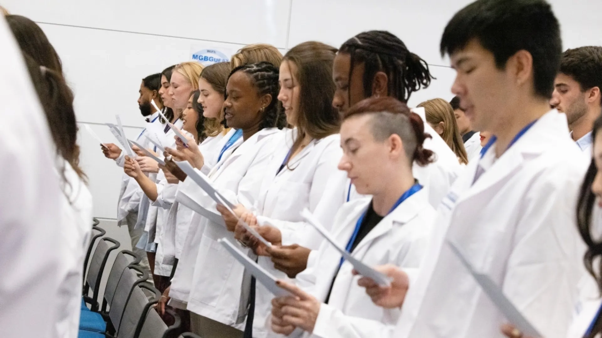 A line of people in white medical coats read from a piece of paper