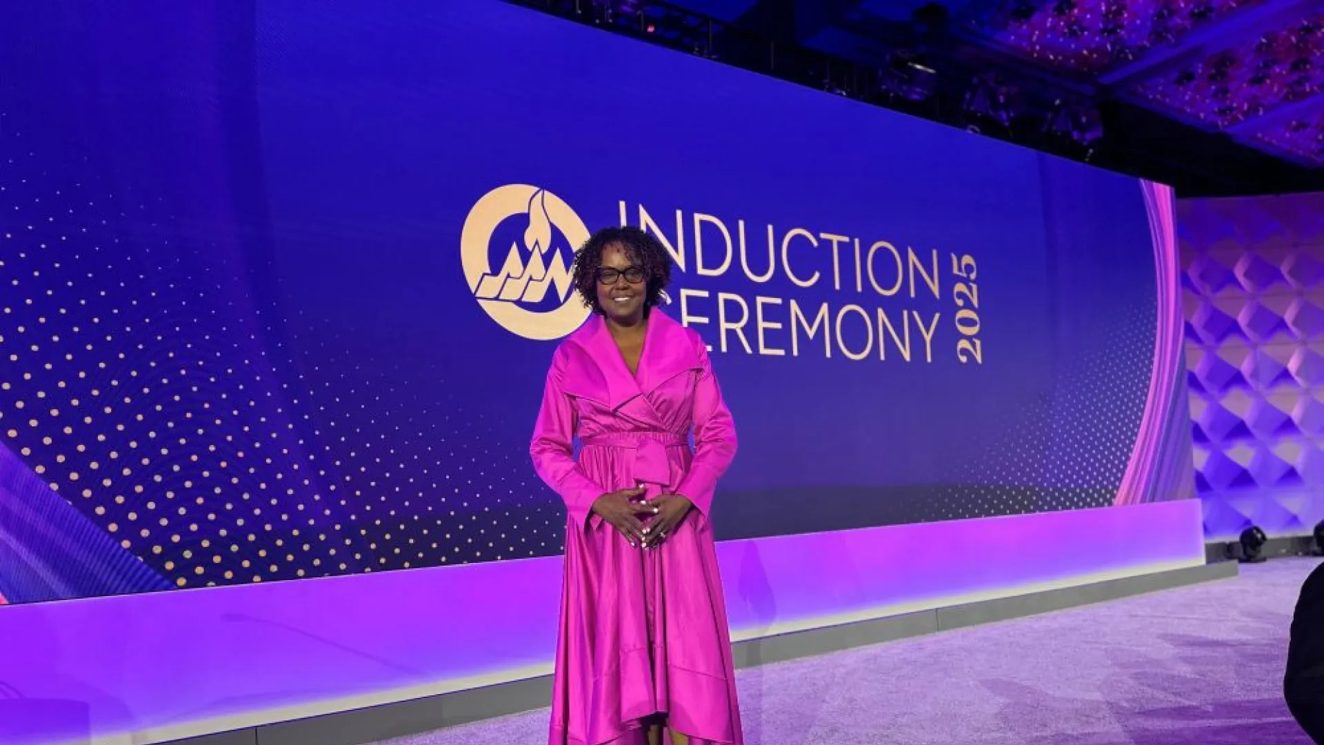 A woman in a pink dress stands on a stage with a backdrop that says Induction Ceremony 2025