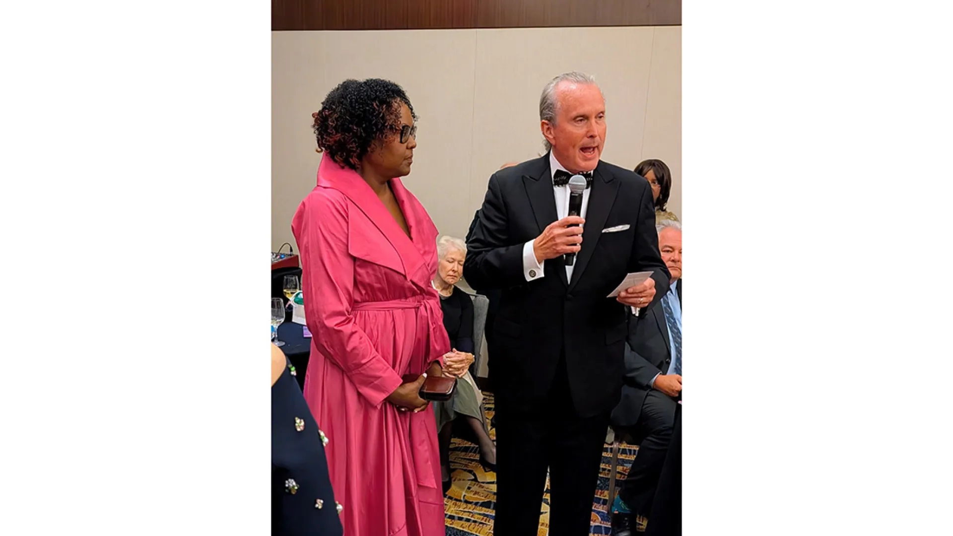 A man with a microphone speaks standing next to woman in pink dress