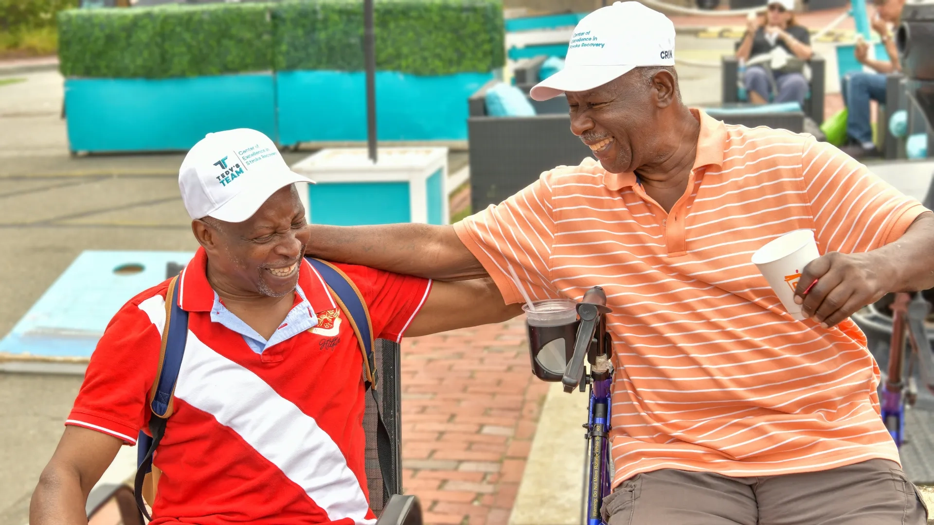 Two men sit outdoors smiling with their arms around their shoulders