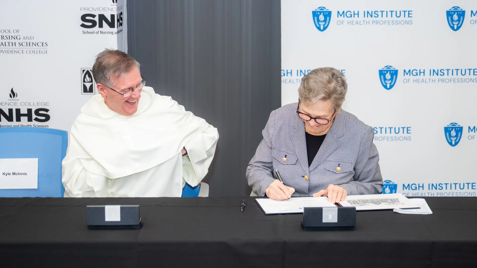 A woman at a table signs a paper while a man looks on