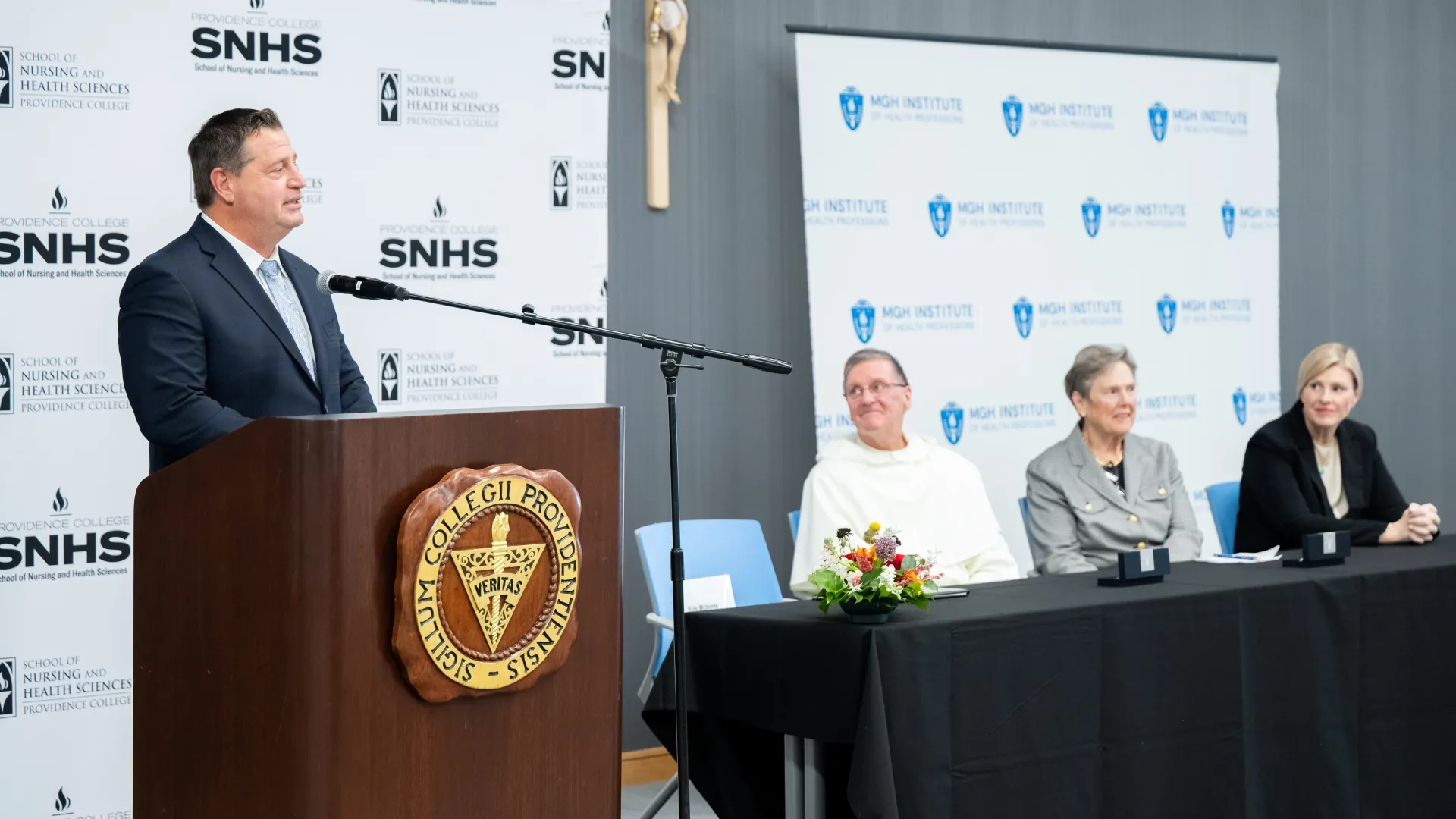 A man stands at a podium while three people sit at a table next to it. 
