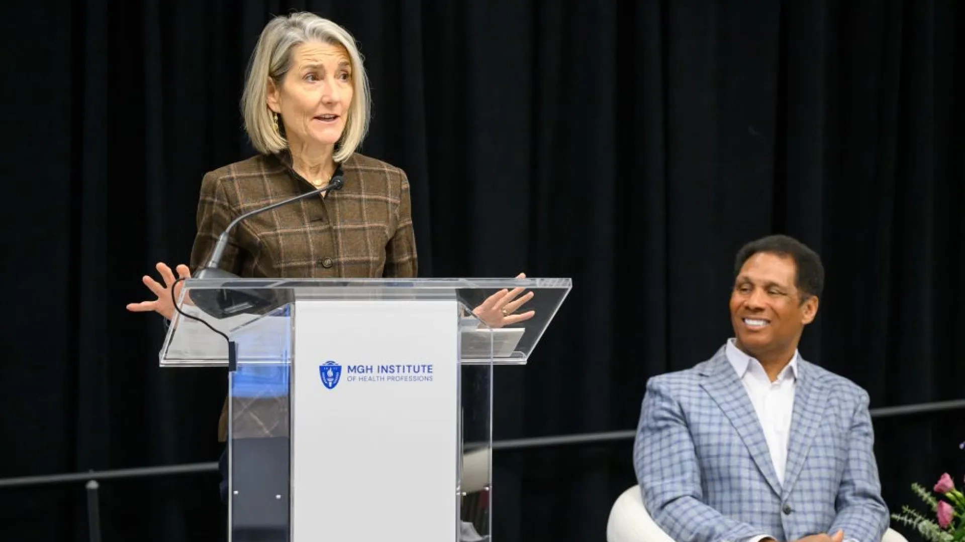 woman standing at podium with man sitting next to her