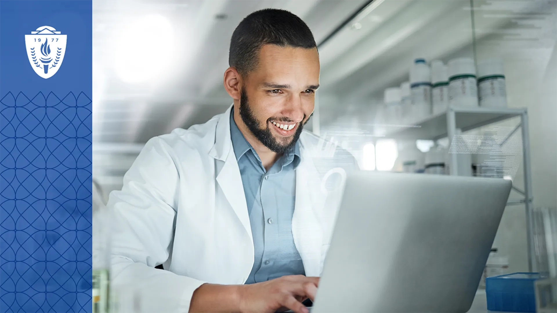 Man wearing blue shirt and white lab coat sitting in a lab and typing on a laptop.